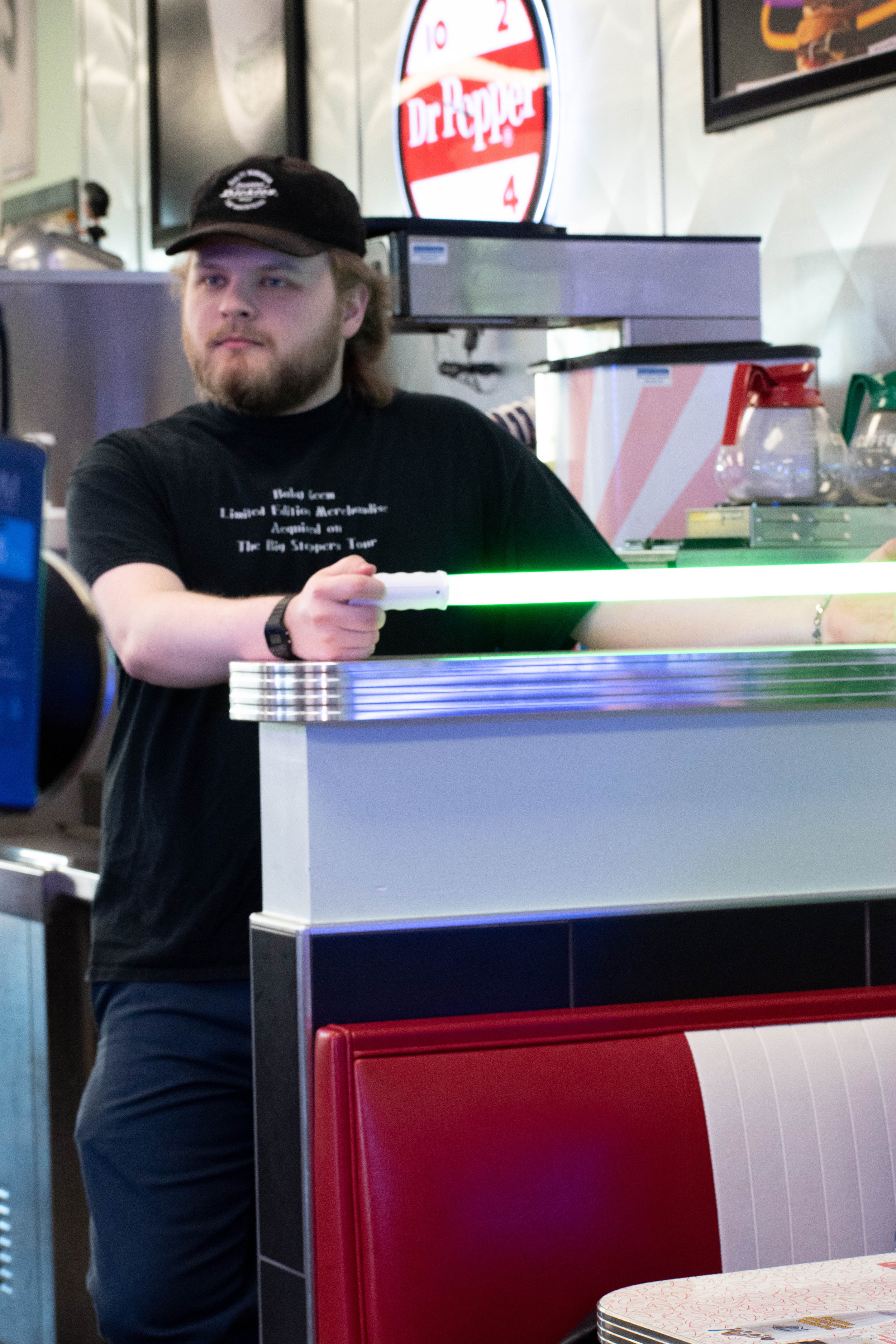 A man with a beard and long hair wearing a black cap and black t-shirt standing behind a counter in a restaurant or cafe, with promotional posters and a Dr. Pepper sign in the background.