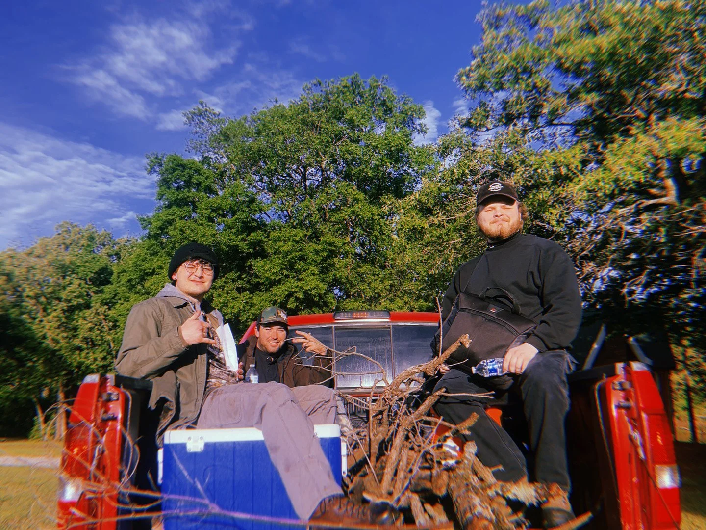 Three men sitting in the back of a red pickup truck outdoors, with green trees and blue sky in the background, holding water bottles and making peace signs.