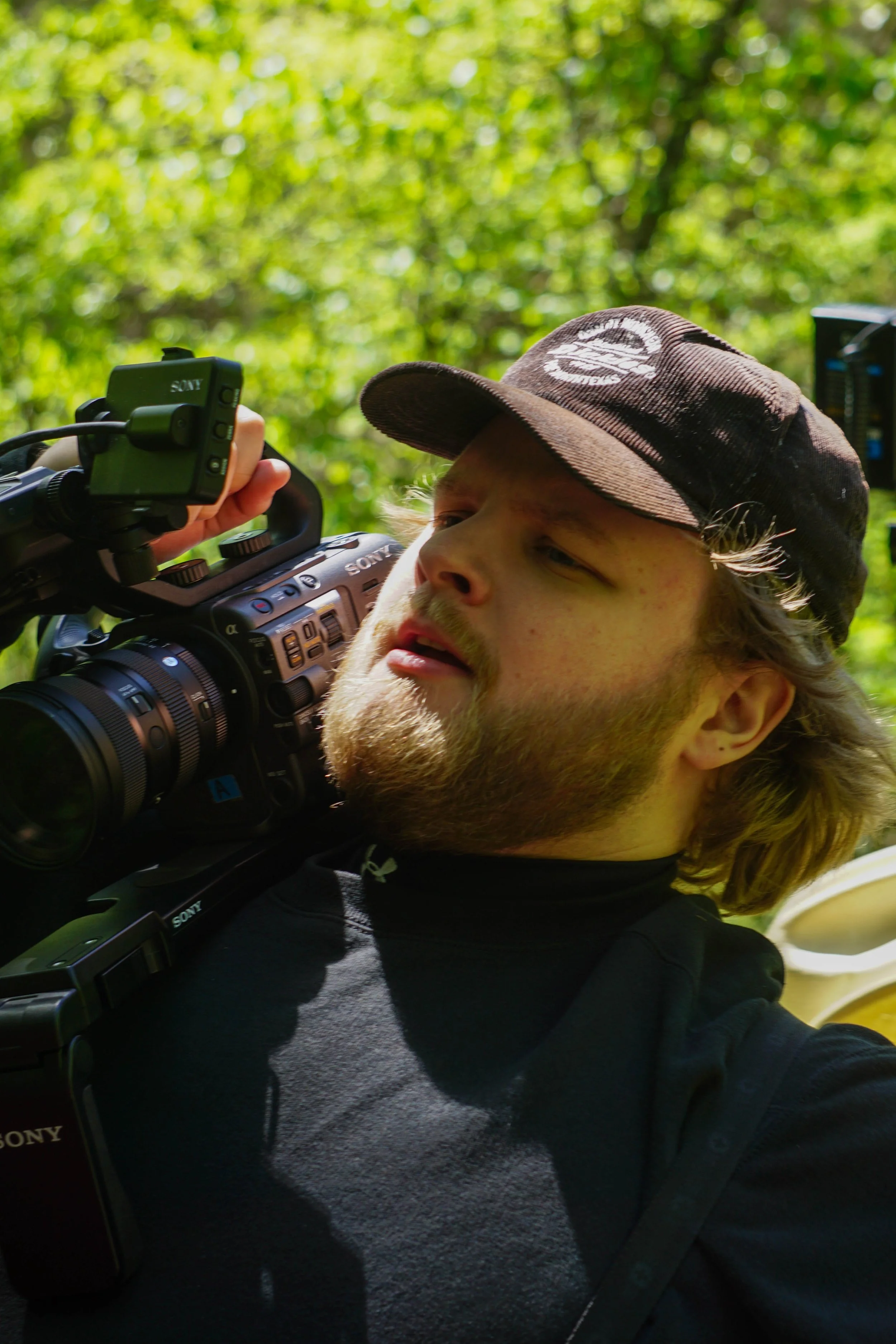 A person with long hair and facial hair, wearing a black shirt and a cap, holding a professional Sony video camera outdoors in a lush green setting.