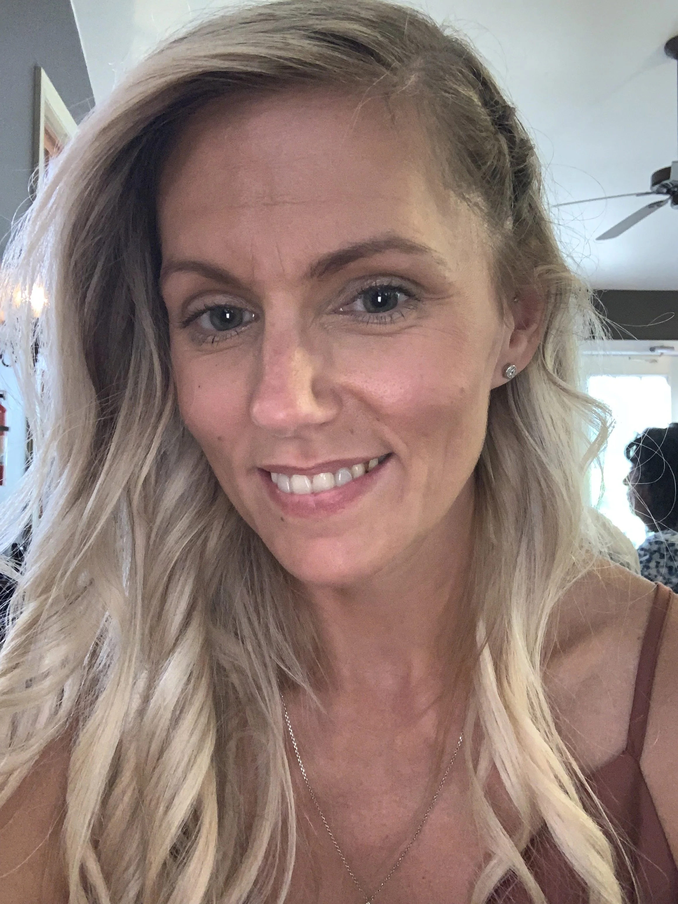 Close-up of a smiling blonde woman with wavy hair, wearing a necklace and earrings, indoors with a ceiling fan in the background.
