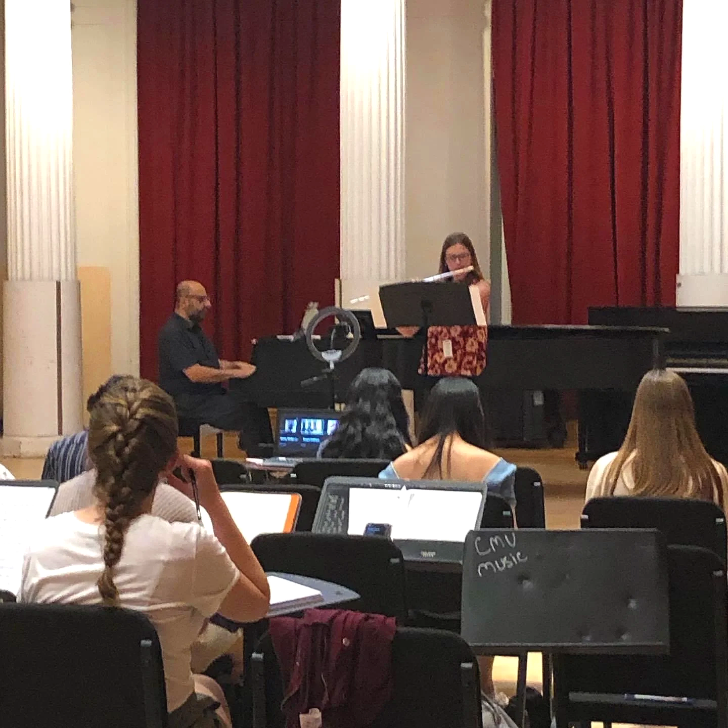 A woman and a man playing the piano on stage during a music recital in an auditorium with red curtains. Audience members are seated in front, some using laptops.