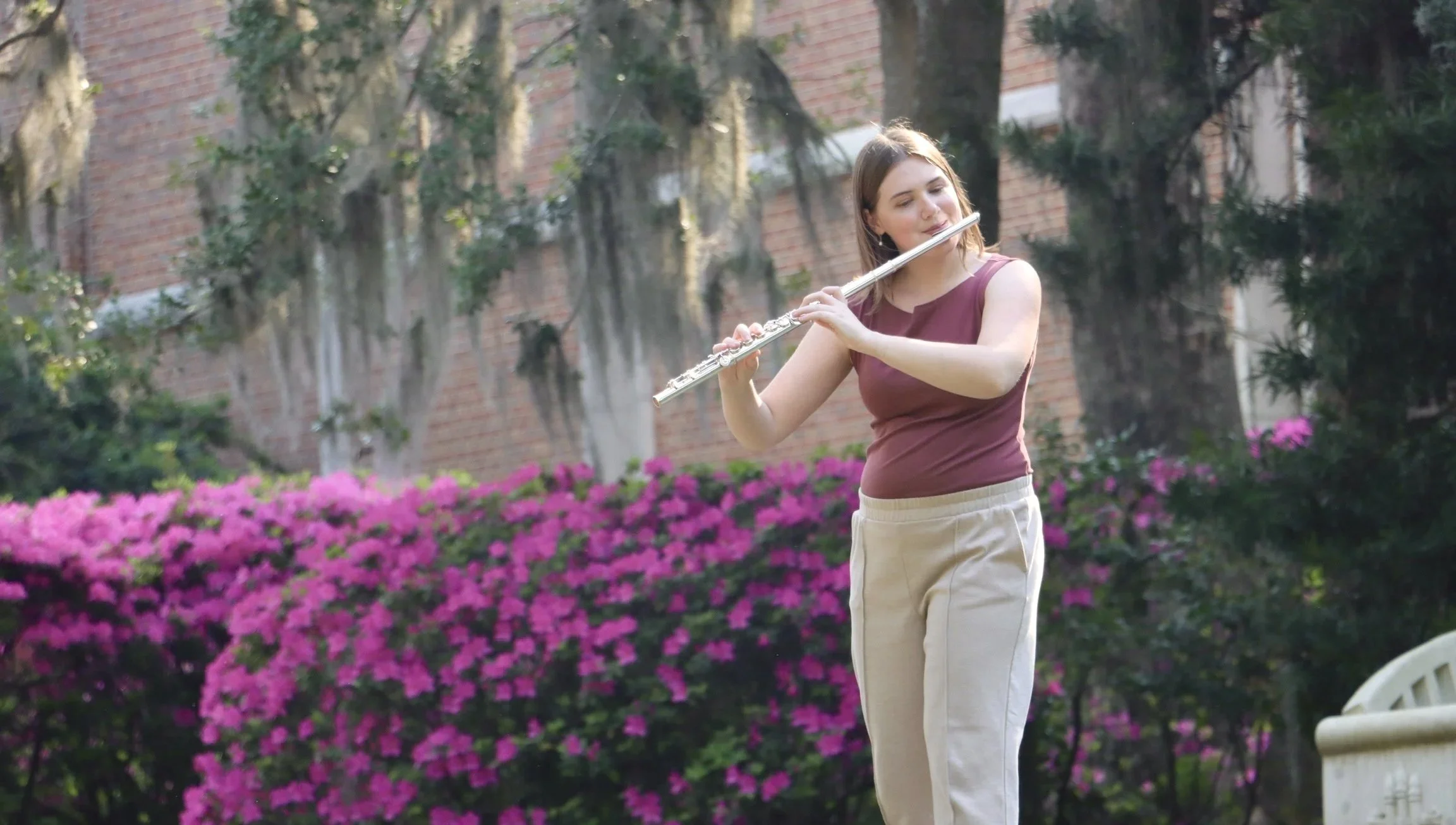 Lindsey Kovach playing the flute outdoors in a garden with pink flowers and green trees.