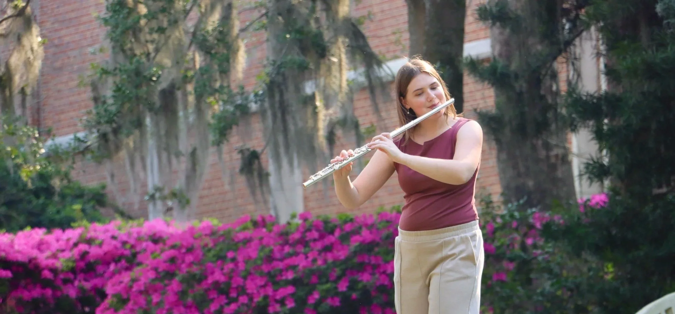 Lindsey Kovach playing the flute outdoors in a garden with pink flowers and green trees.