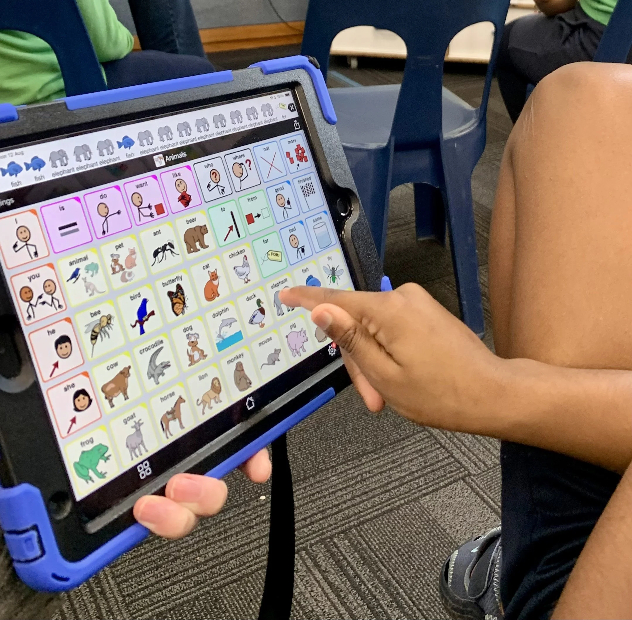 Close-up of a person holding a tablet displaying an educational app with pictures of animals and words, with other children seated nearby.