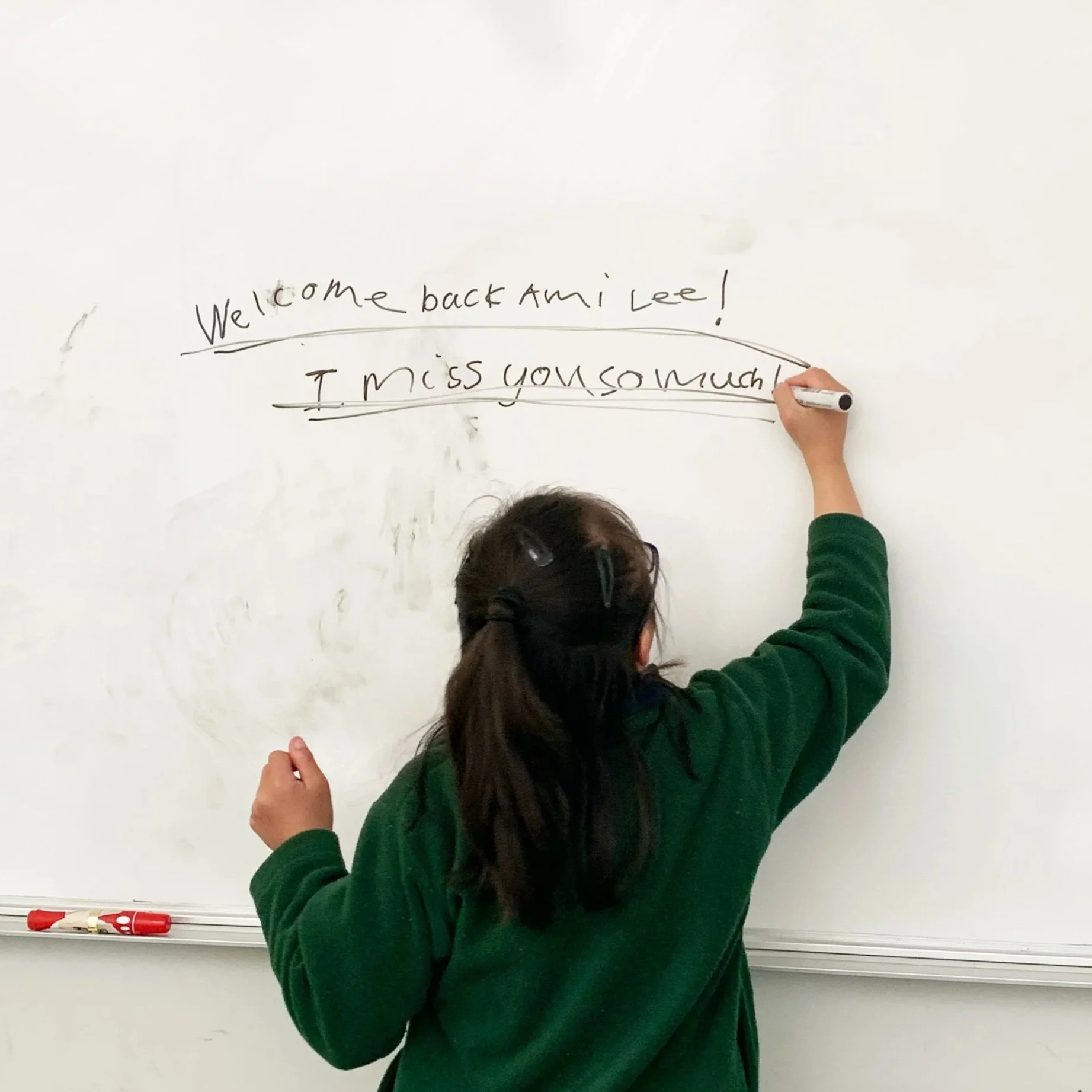 Person writing on a whiteboard with markers, with the words "Welcome back Ami Lee! I miss you so much".