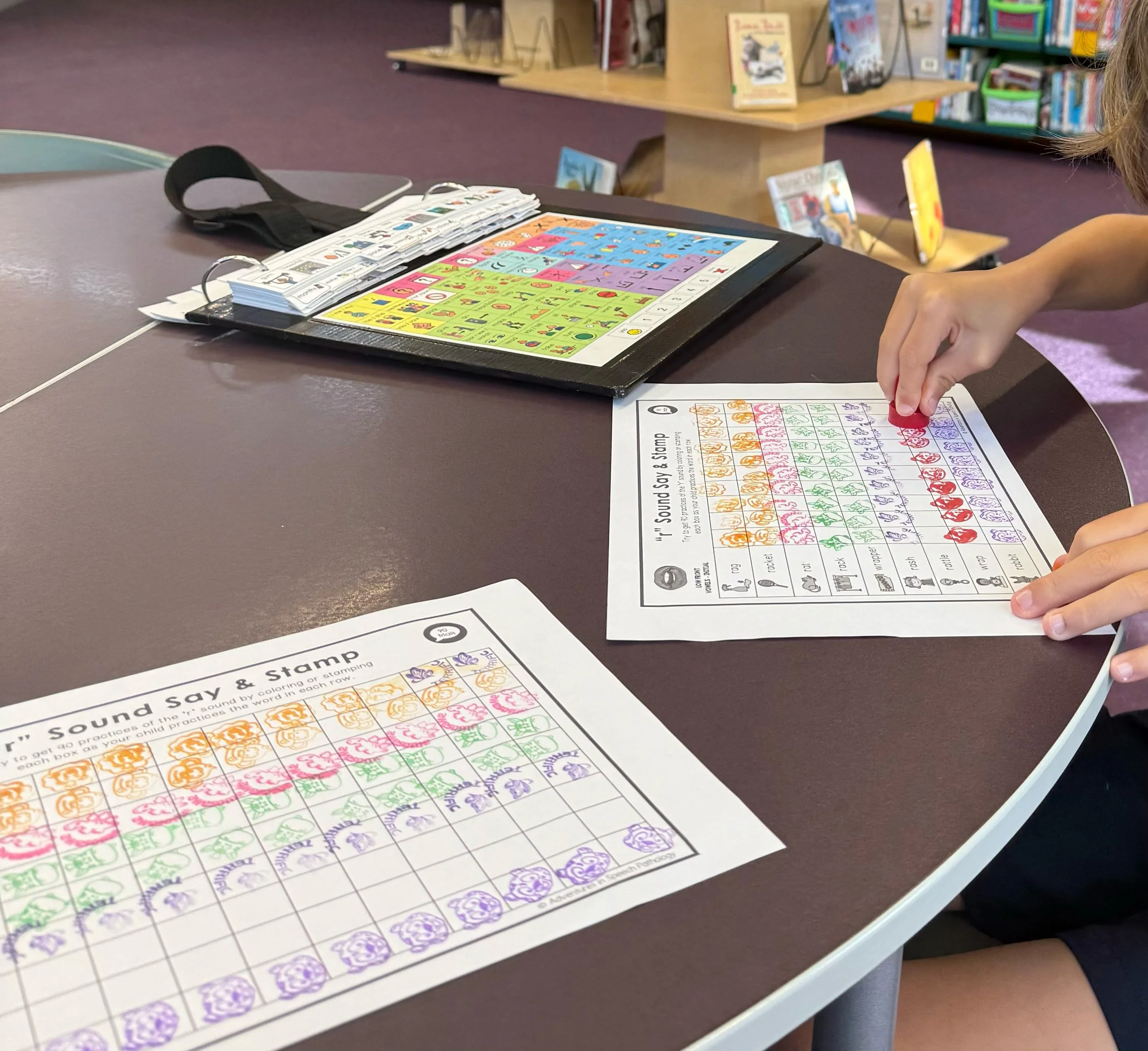 A child's hand stamping a colorful alphabet and animal-themed worksheet on a table in a library or classroom setting. In the background, there are bookshelves filled with children's books and a wooden table with small display stands holding books.