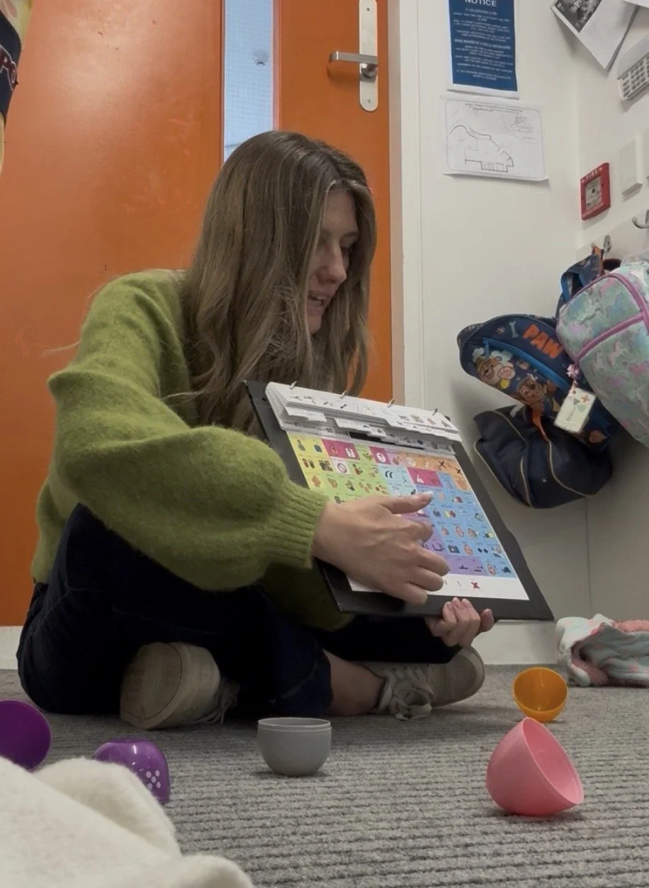A woman sitting on the floor, reading a colorful book or pamphlet, surrounded by children's toys and backpacks in a room with an orange door and various notices on the wall.