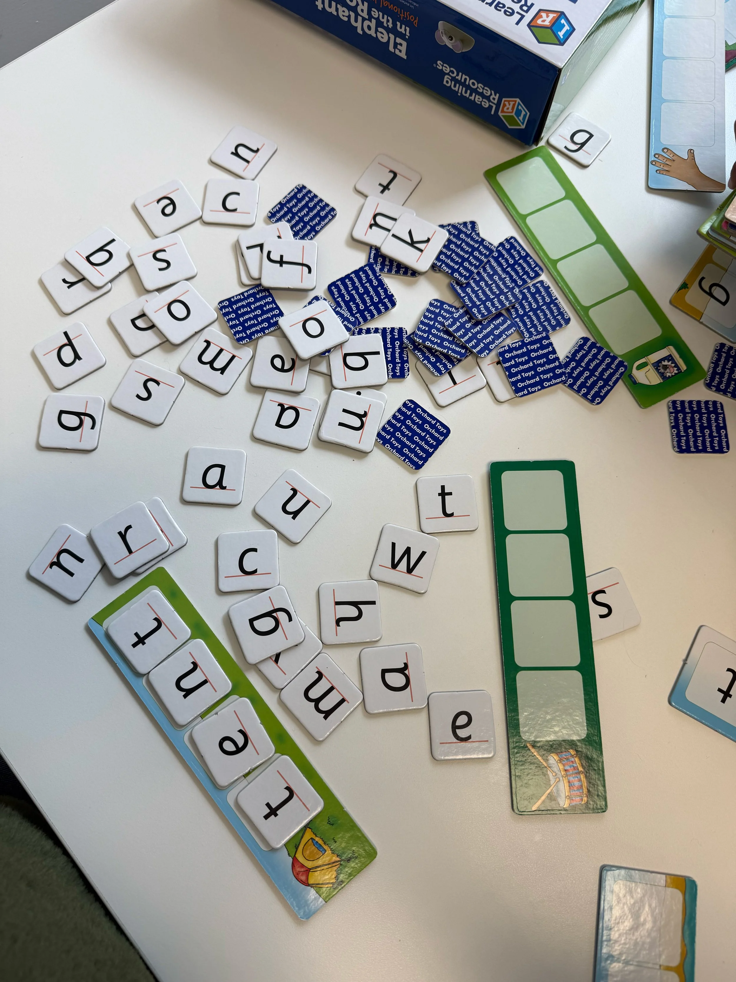 A tabletop with scattered letter tiles, game cards, and a game board. The letter tiles display various lowercase letters, some arranged and some scattered. The game cards have pictures and are from a game called Leapster Learning in English. The game box is partially visible, showing colorful text and graphics.