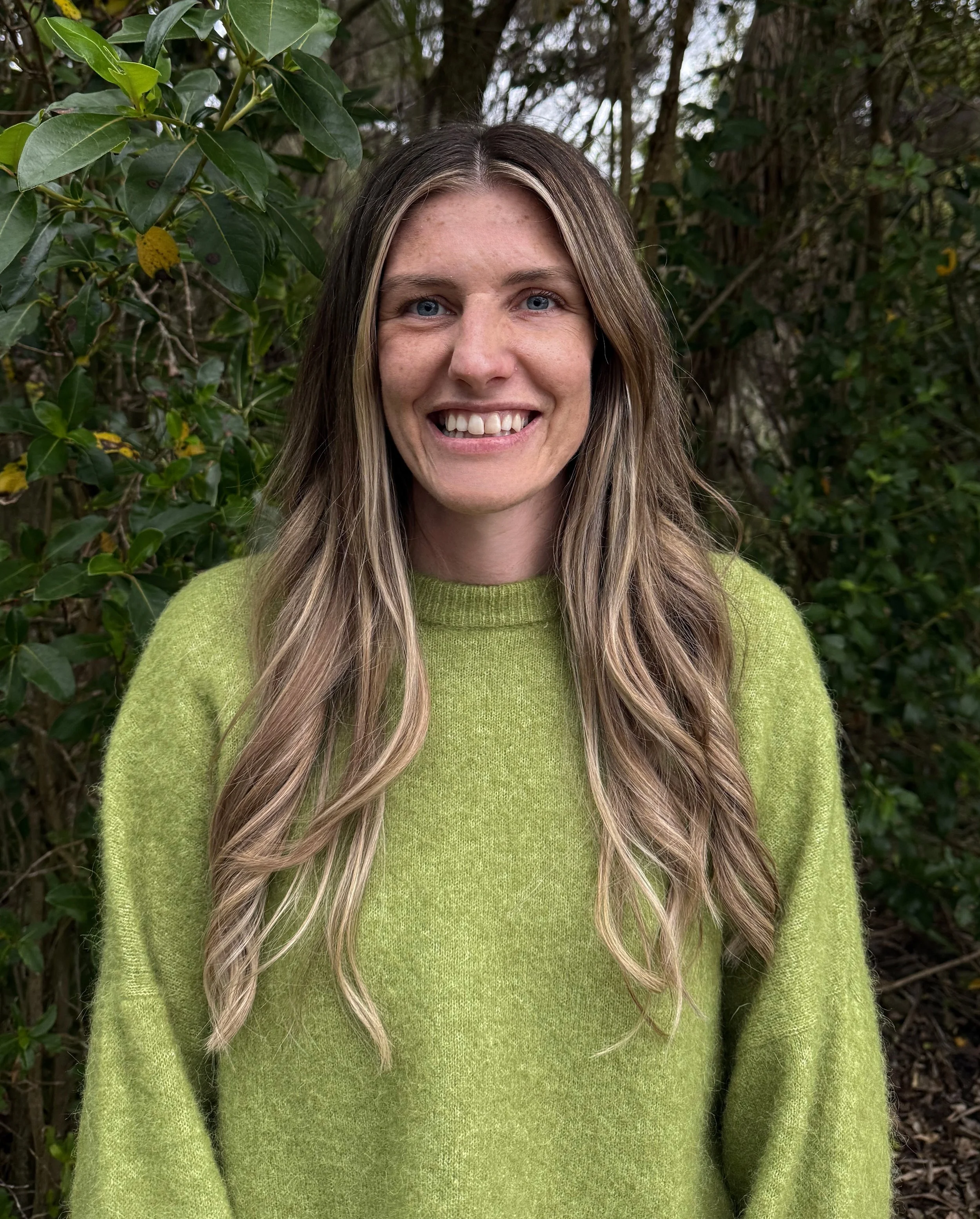 A woman with long, wavy hair and blue eyes smiling outdoors in front of green foliage, wearing a bright green sweater.