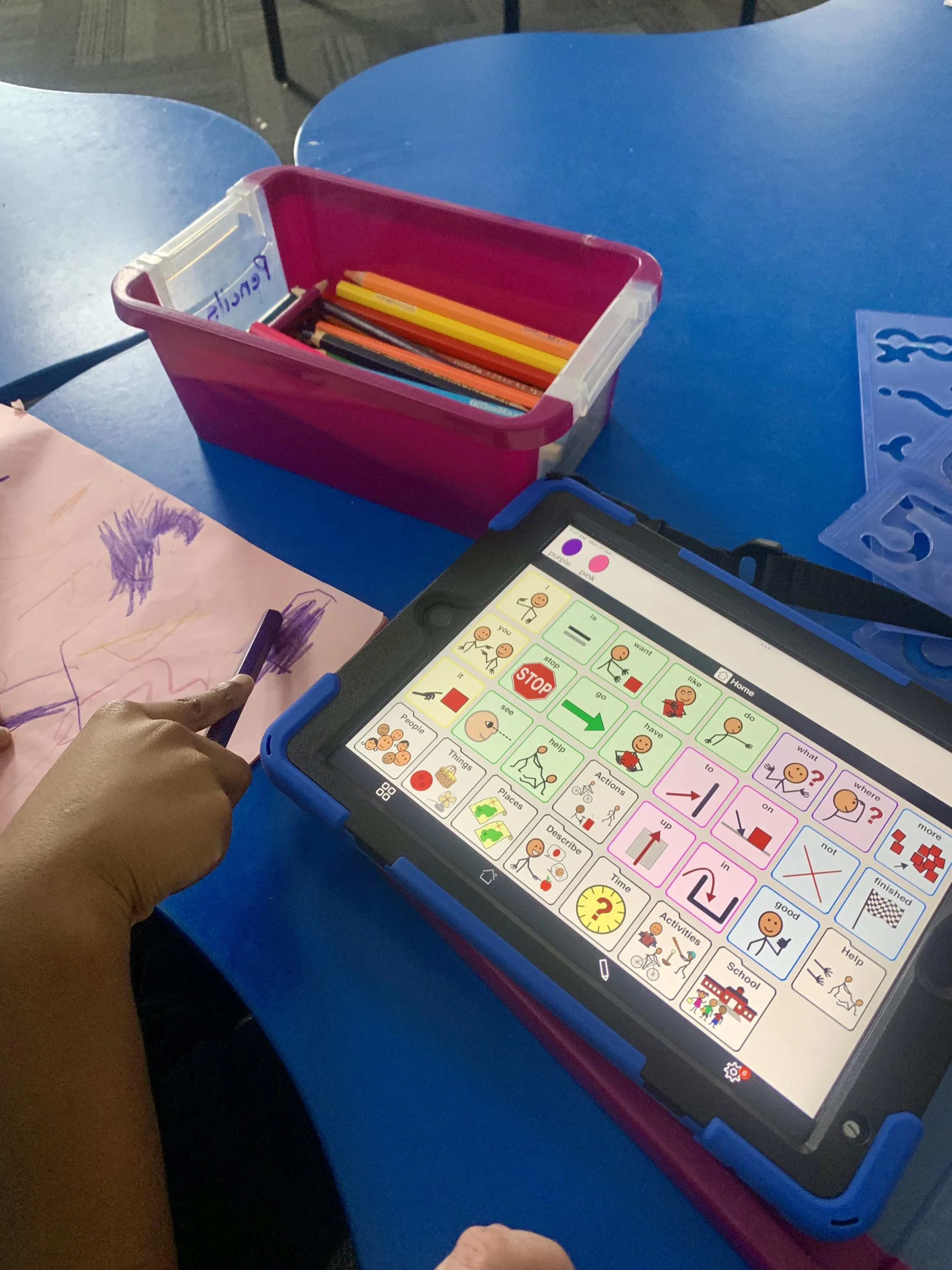 Tablet with communication symbols and picture icons on a bright screen on a blue desk, with a pink container holding colorful pencils nearby.