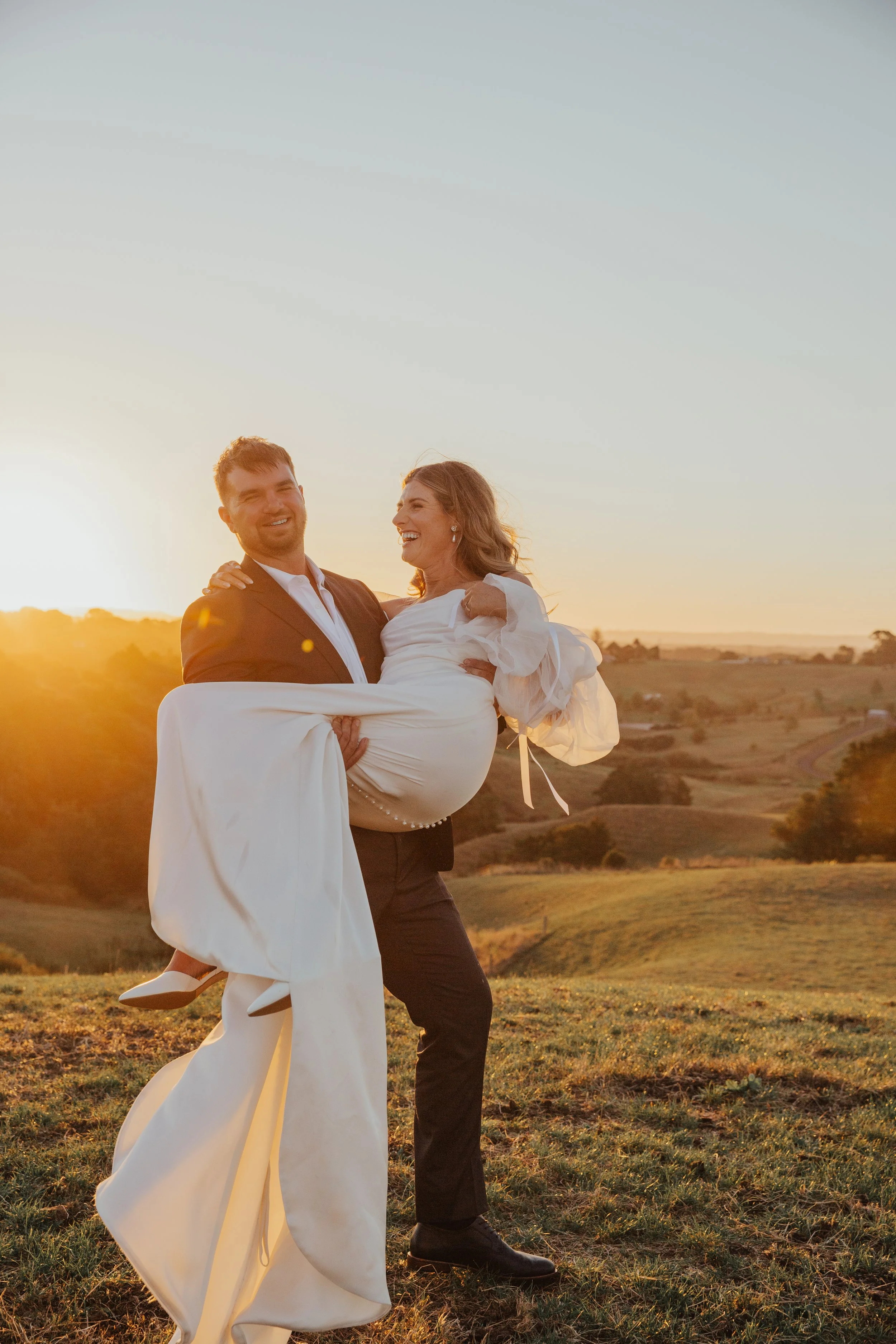 A couple dressed in formal attire outdoors during sunset. The man is holding the woman in his arms, and they are smiling and laughing. The background features rolling hills and a clear sky with warm sunlight.