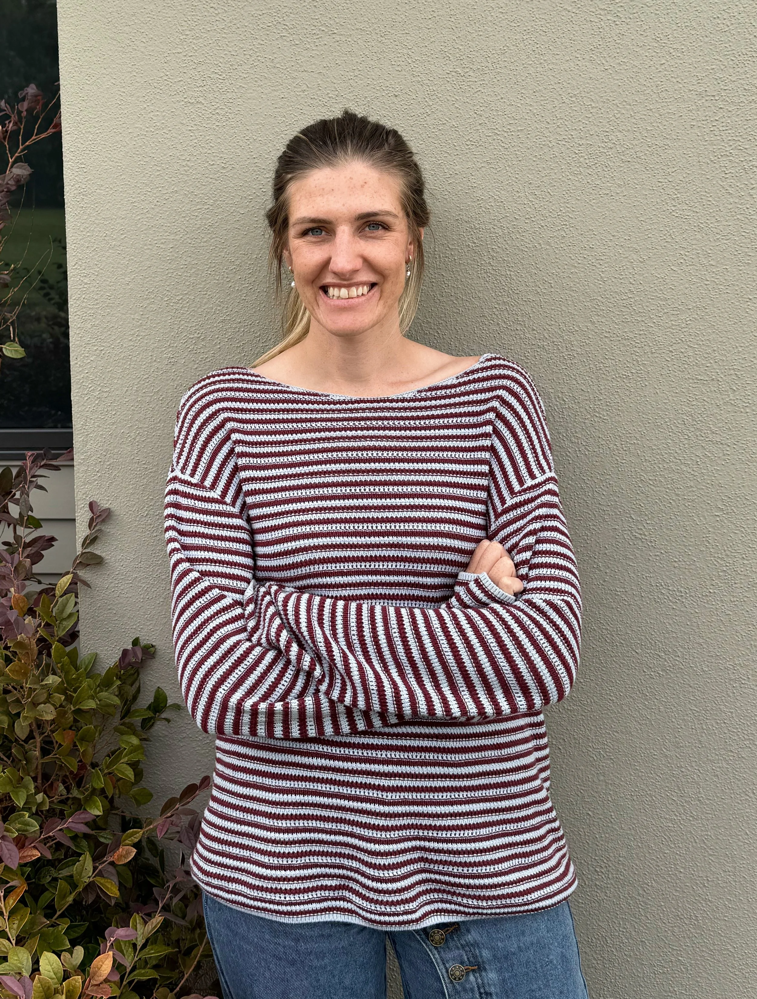 A woman standing outdoors in front of a beige textured wall, wearing a striped long-sleeve shirt and blue jeans, smiling with arms crossed.