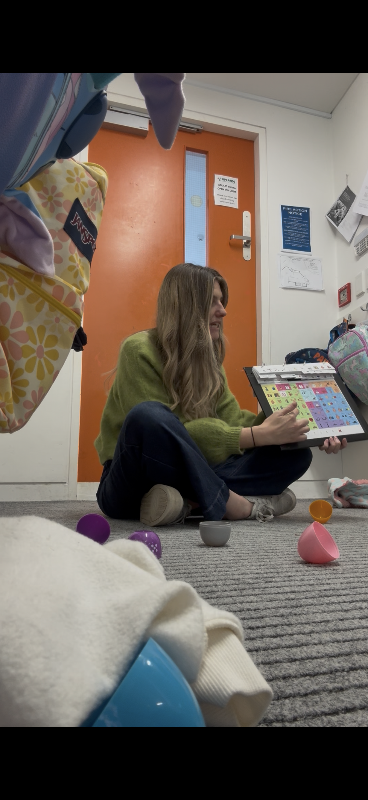 A young woman with long wavy hair, sitting cross-legged on the floor, holding a picture book with colorful illustrations. She is wearing a green sweater and dark pants, and appears to be in a waiting area or hallway, with a closed orange door behind her and various supplies and toys around her.