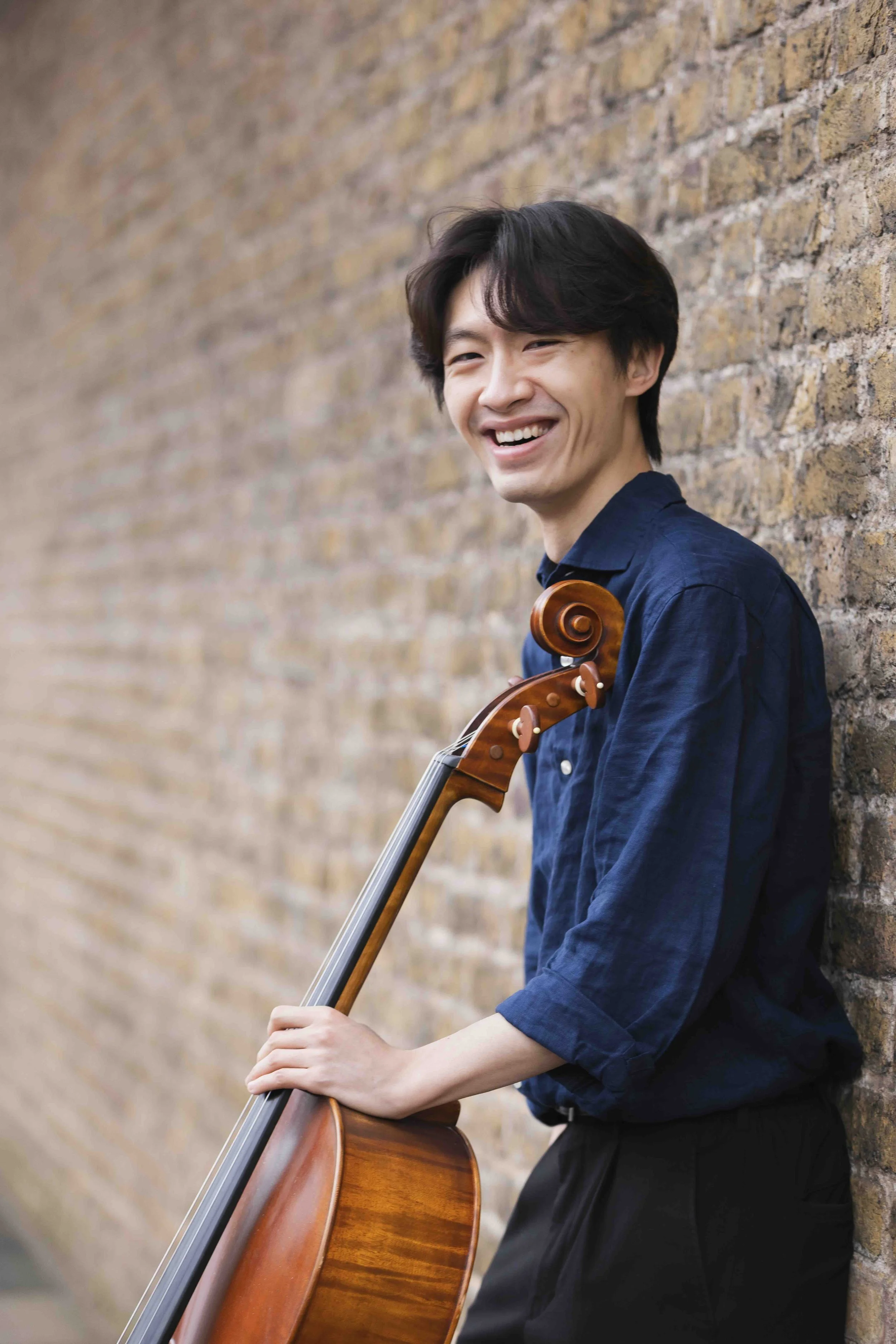 A man with dark hair and a joyful smile, wearing a navy blue shirt, playing a wooden cello outdoors against a brick wall.