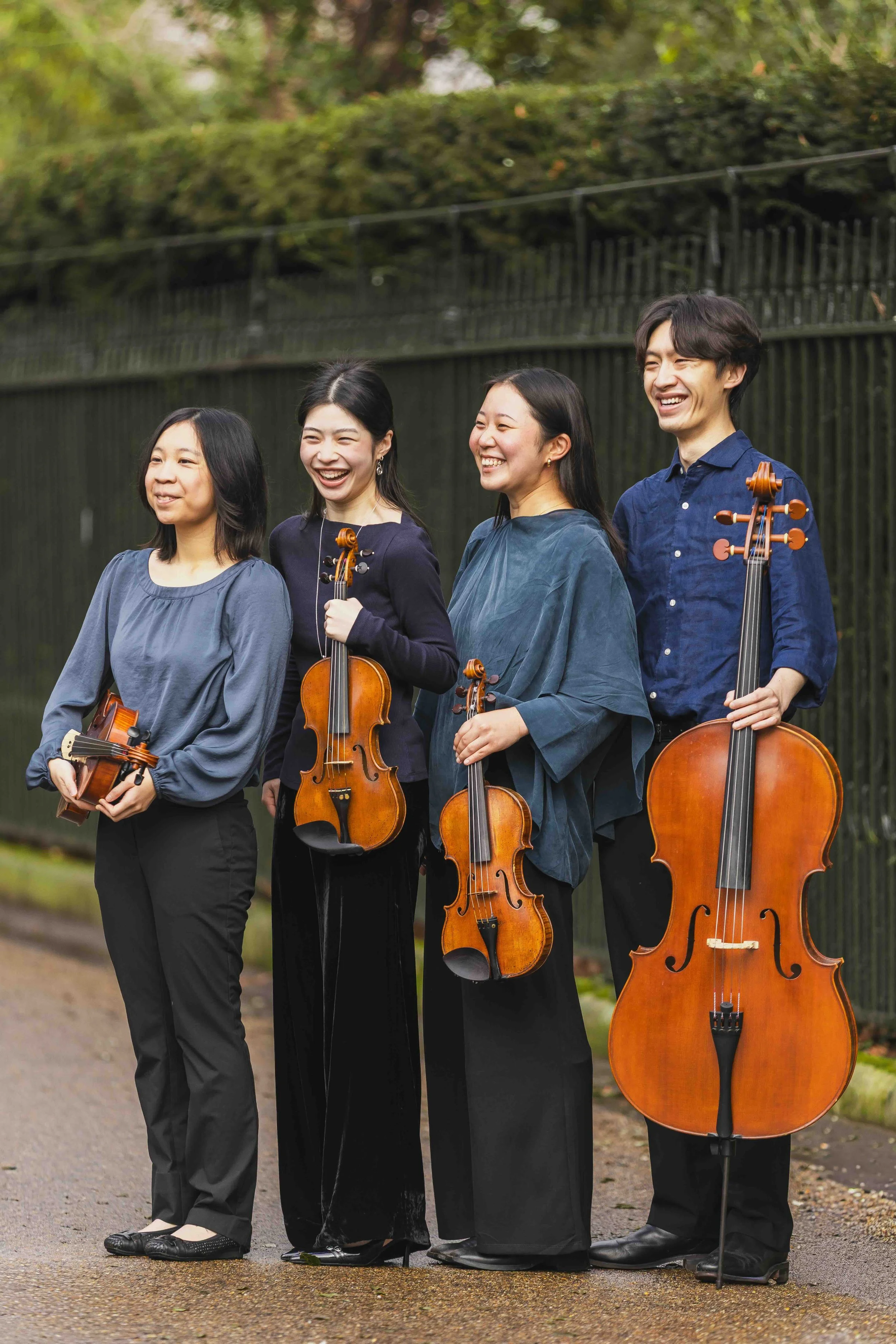 Four musicians holding violins and a cello standing outdoors on a gravel path in front of a black fence and green trees, smiling and laughing.