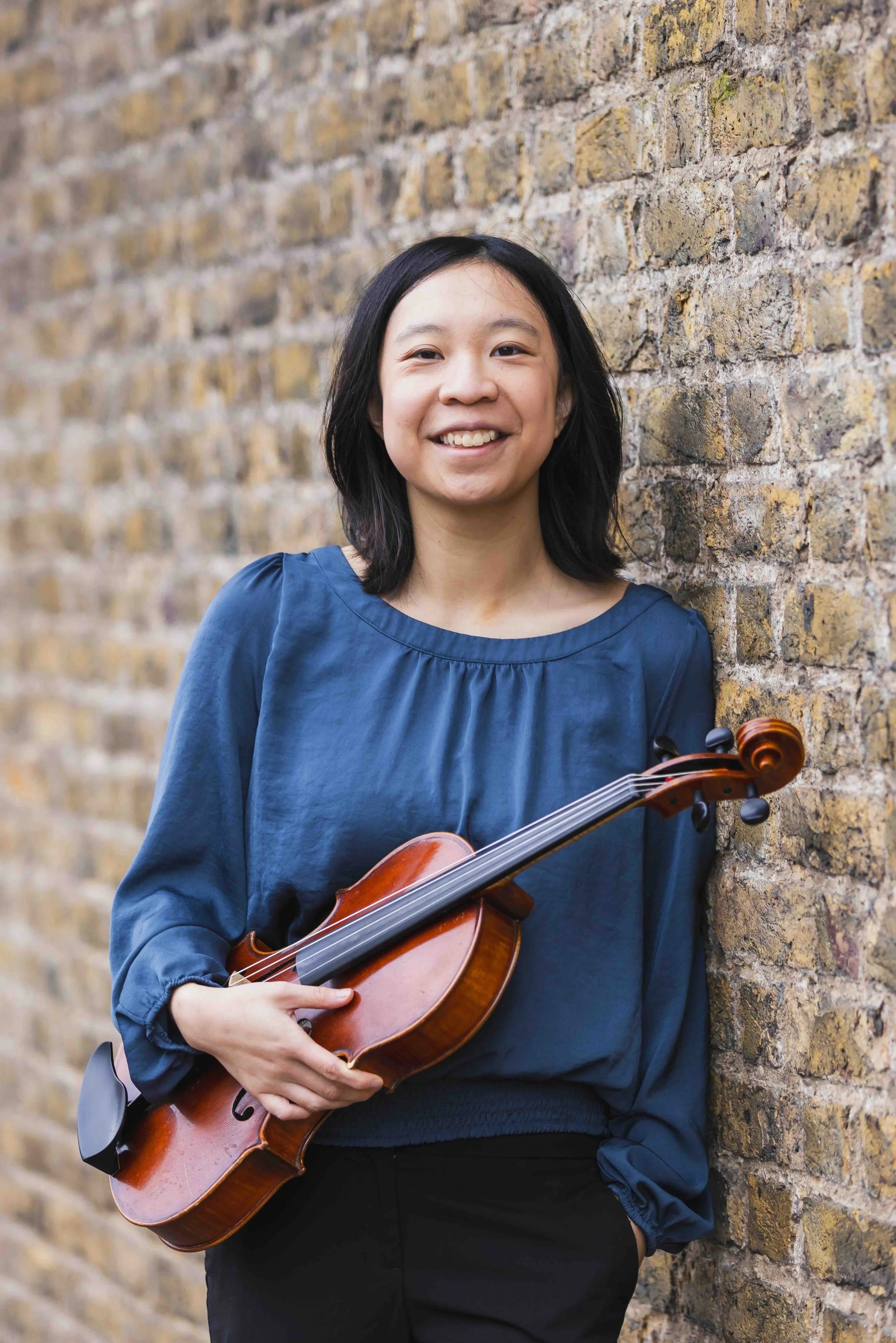 A young woman with shoulder-length black hair smiling while holding a violin, standing against a brick wall.