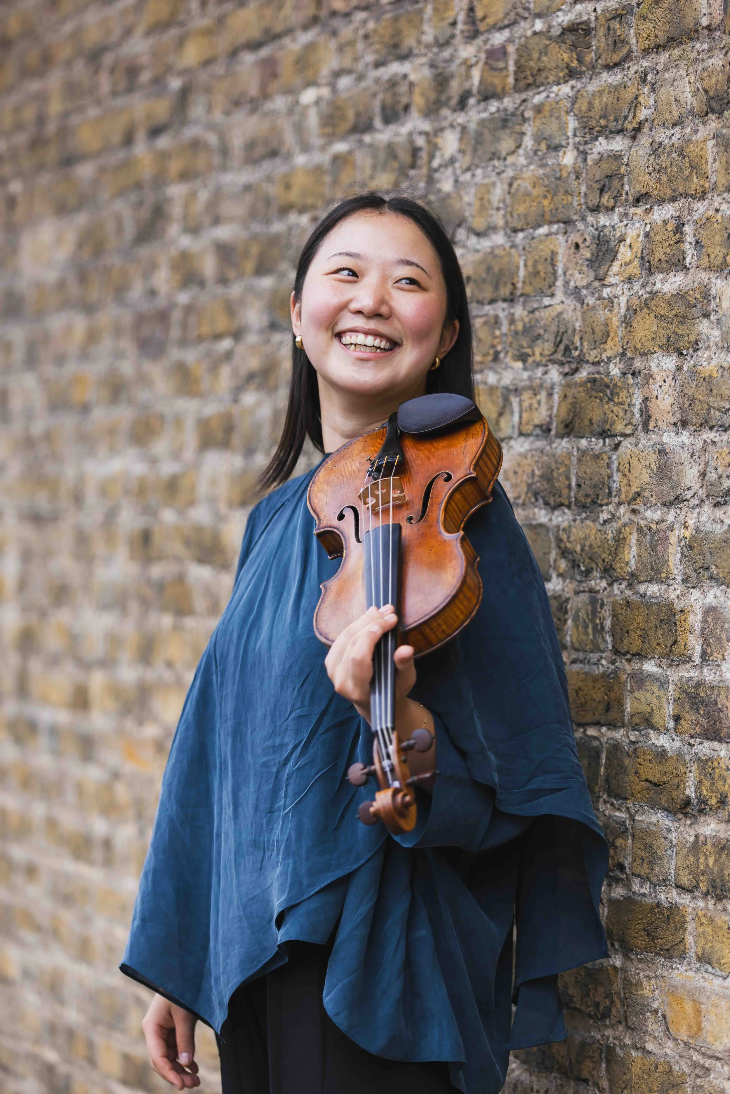 A woman smiling while holding a violin, standing against a brick wall.