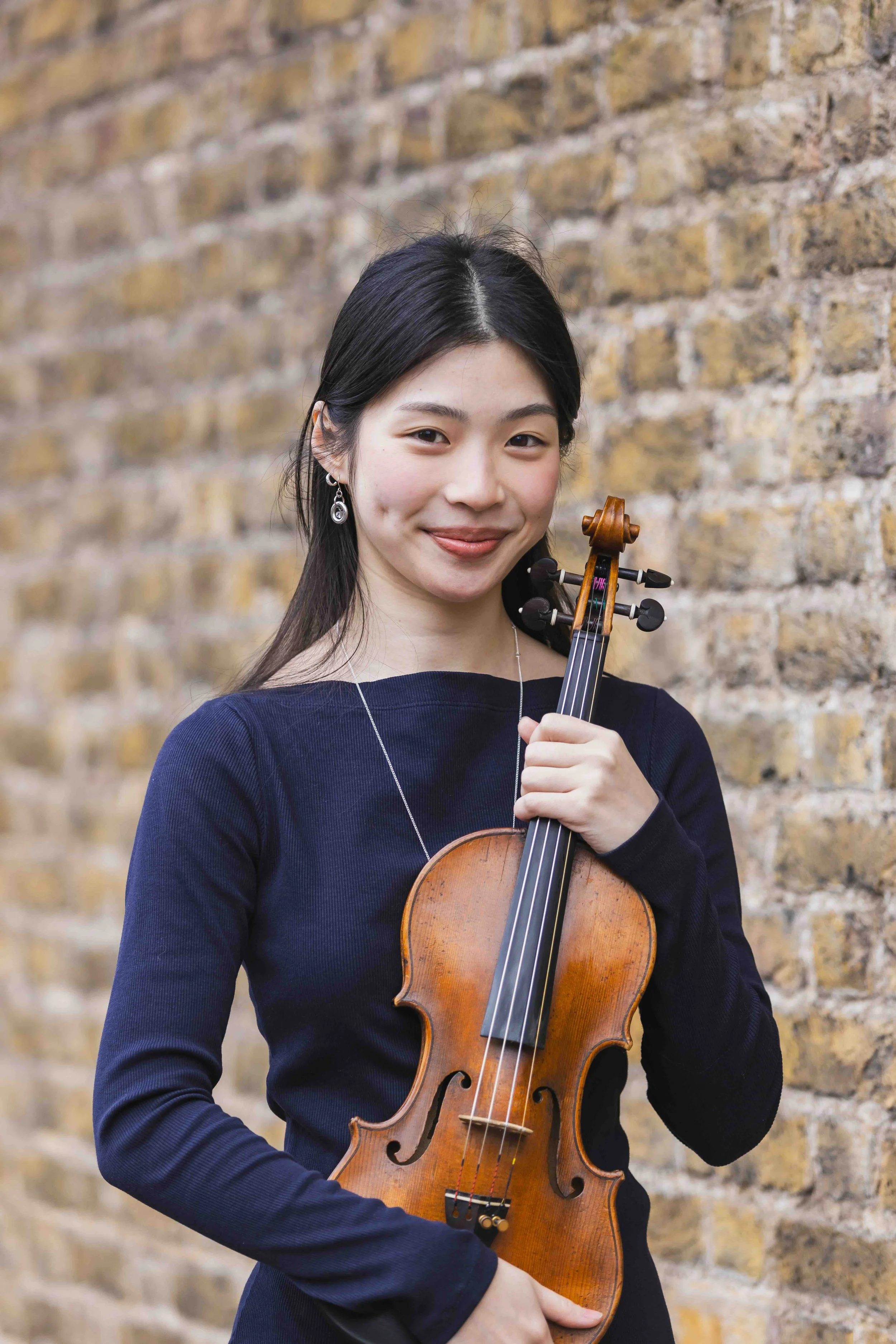 A woman with long black hair and earrings holding a violin against a brick wall