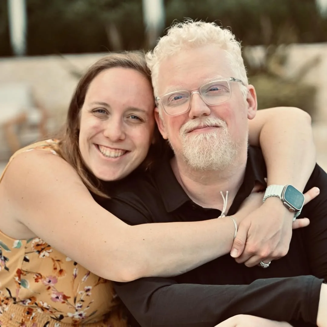 A smiling woman with brown hair hugging a man with white hair, glasses, and a white beard, both looking at the camera outdoors.