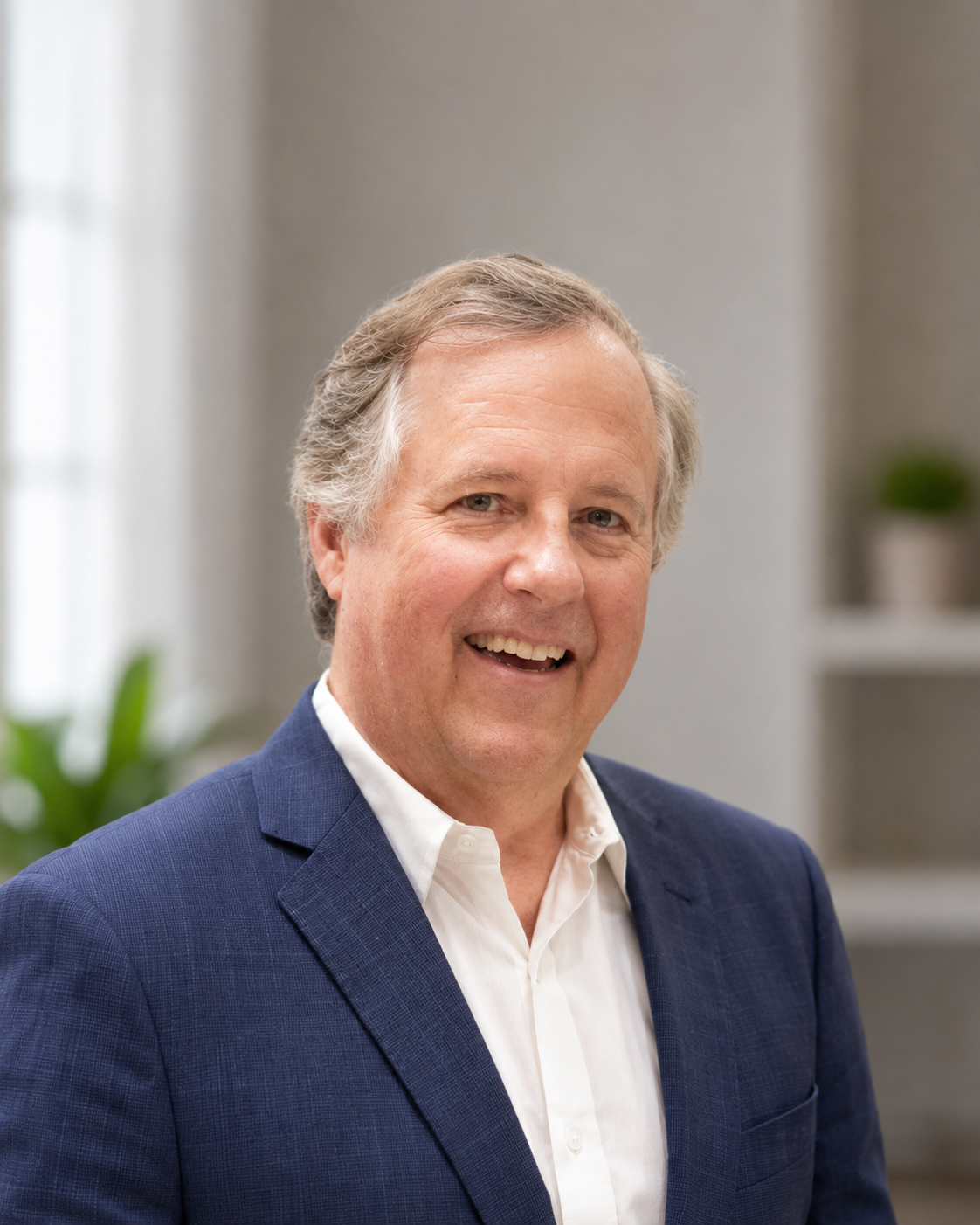 A middle-aged man with gray hair smiling, dressed in a white shirt and navy blue blazer, posing in an indoor setting with blurred background and green plants.
