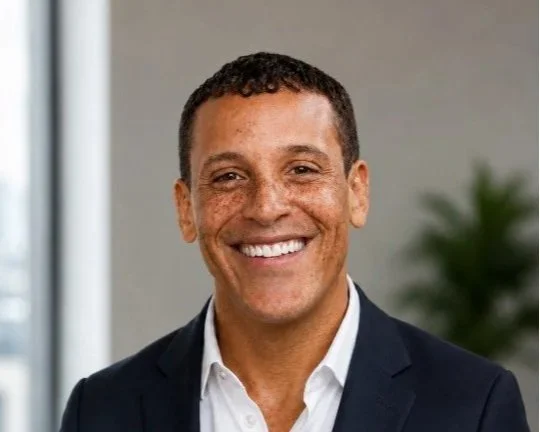 A smiling man with short dark hair and freckles, wearing a dark button-up shirt, standing indoors with a blurred background and soft lighting.