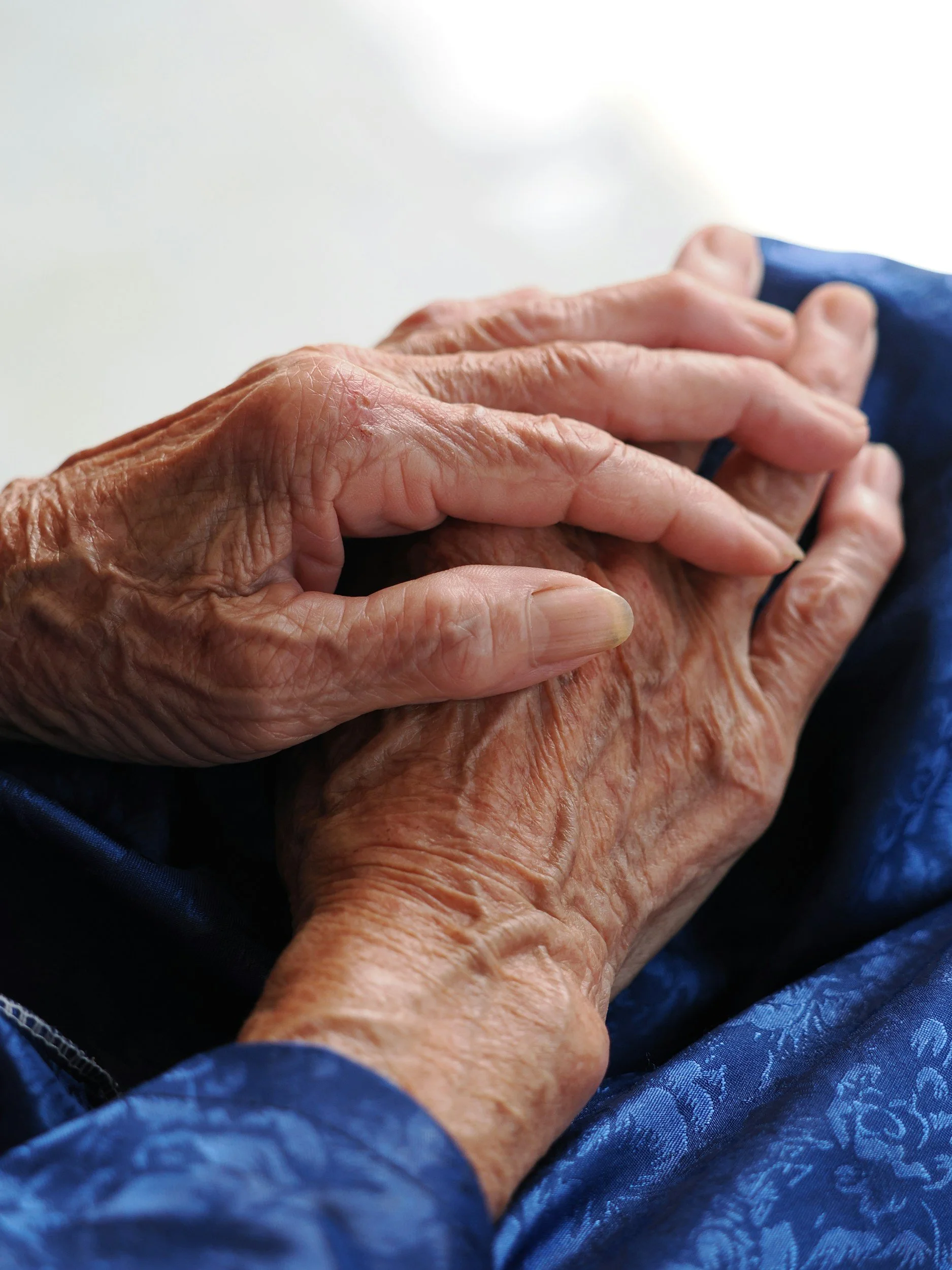 Close-up of two elderly hands holding each other, one hand with prominent wrinkles and age spots, resting on a person's lap covered with blue patterned fabric.