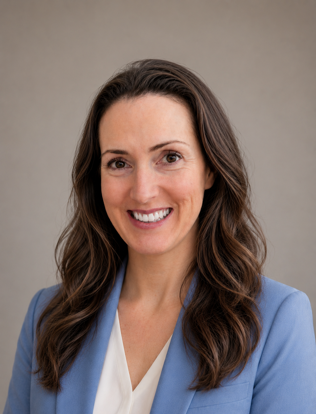 A woman with wavy brown hair, smiling, wearing a light blue blazer and white blouse, against a plain gray background.
