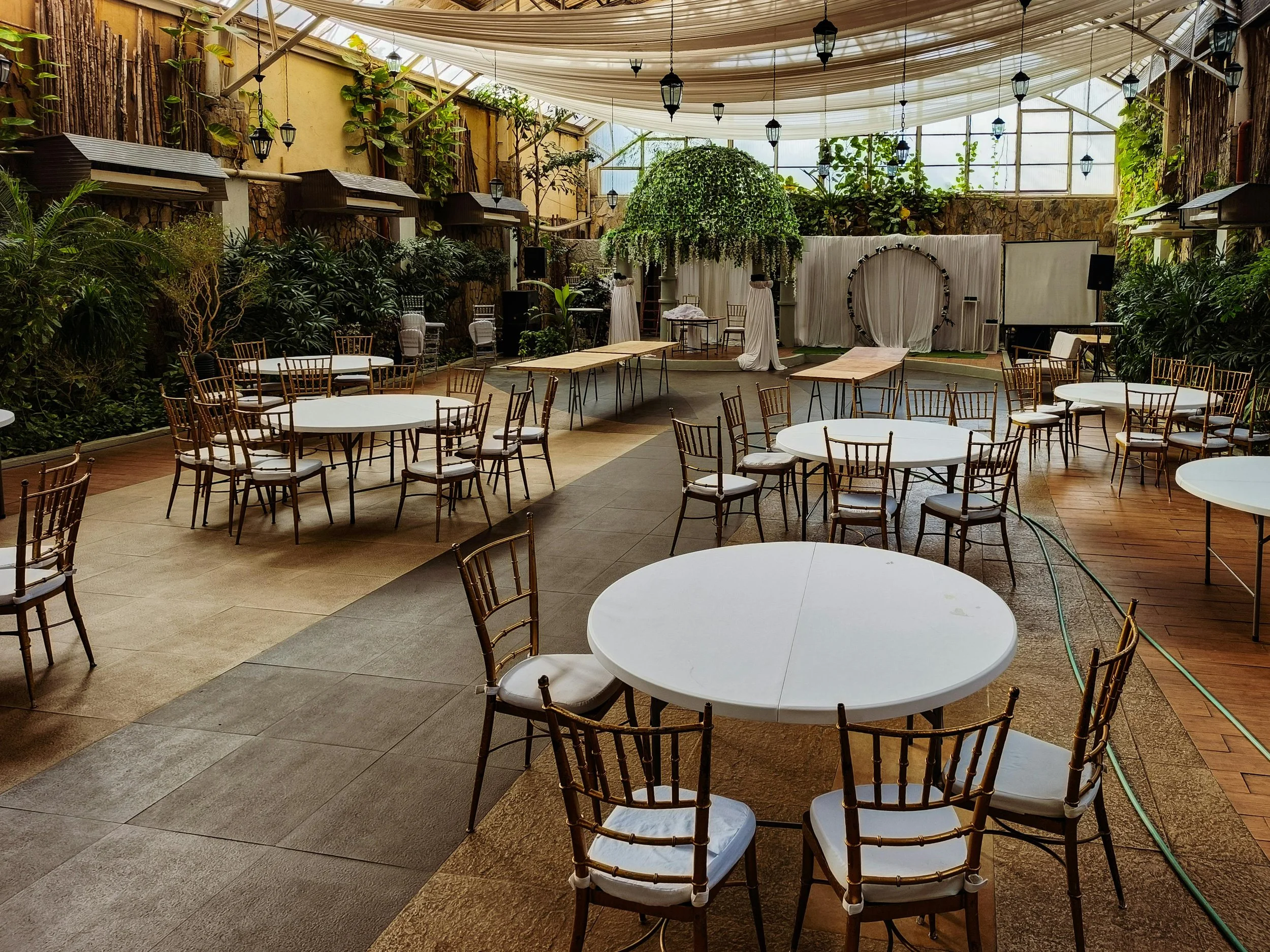 An indoor garden venue decorated for an event with round tables and chairs, lush green plants, hanging lanterns, draped fabric ceiling, and a small stage with a white curtain backdrop.