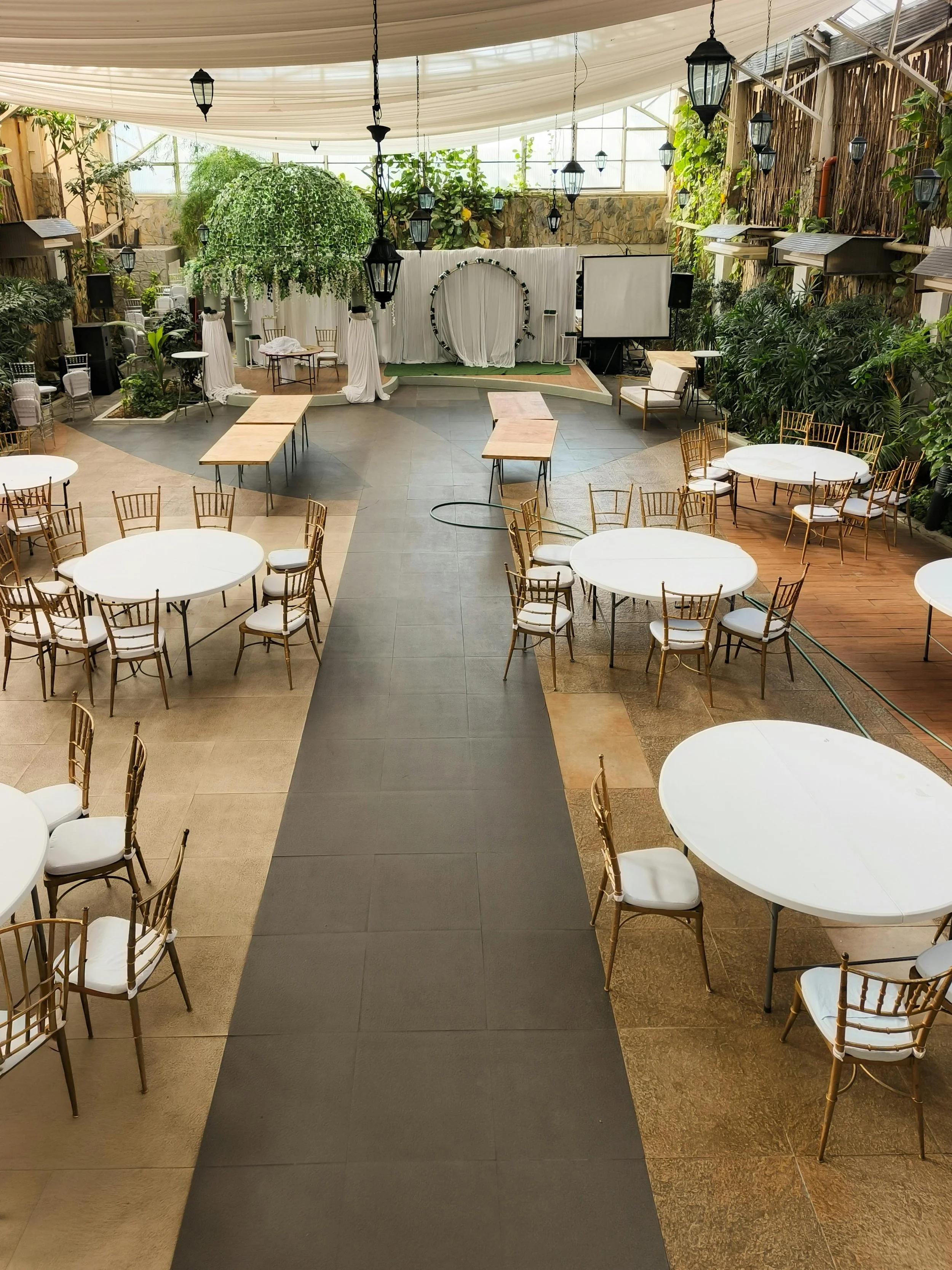 An indoor event space set up for a wedding or celebration, with round banquet tables and gold chairs, a stage with white draping, greenery, and hanging lanterns.