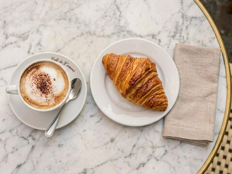 A cup of coffee with foam and a croissant on a white plate on a marble table, with a napkin on the right.