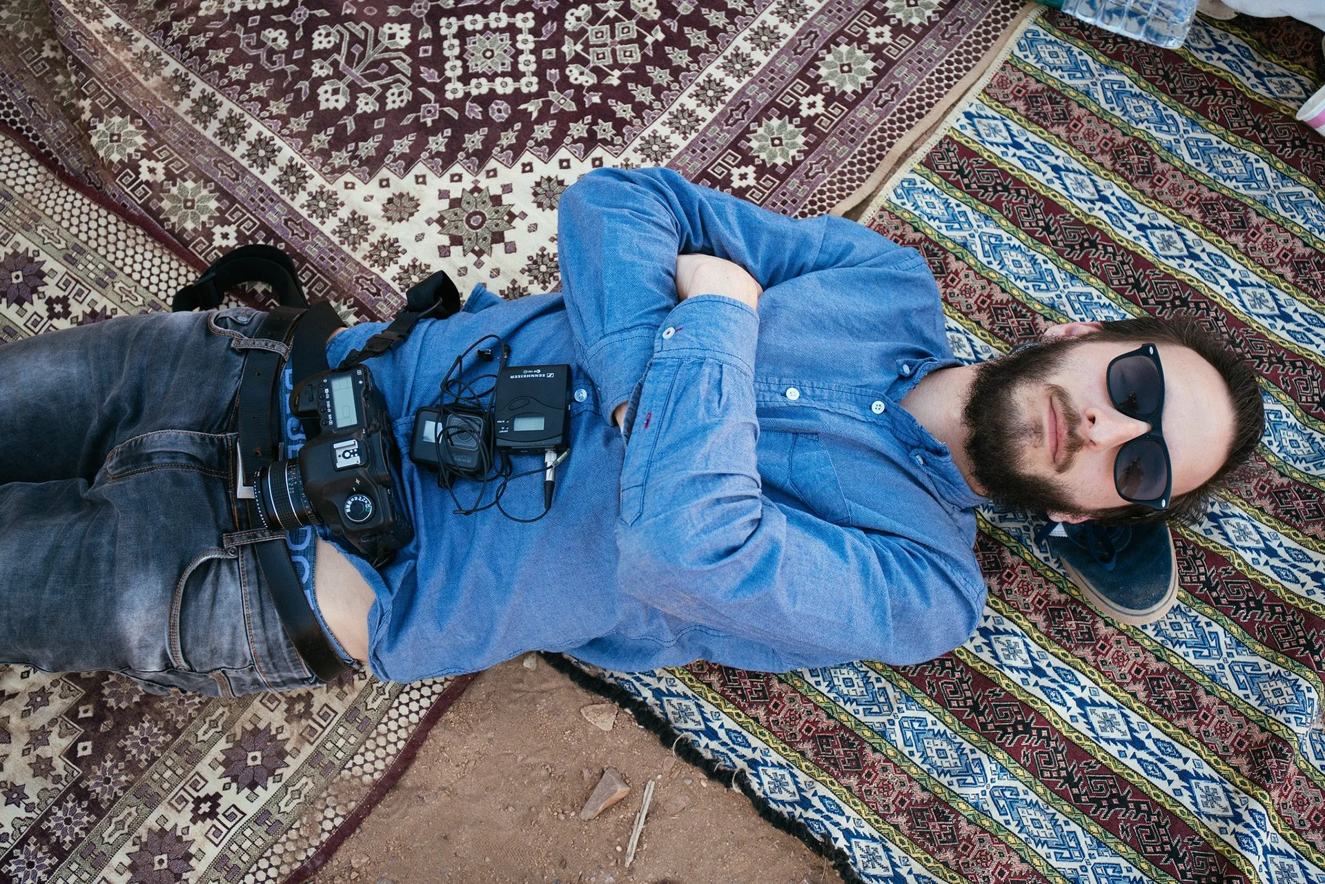 A man with Dark hair and a beard, wearing sunglasses, a blue denim shirt, and jeans, is lying down on a patterned colorful rug outdoors. He has a camera and other electronic devices attached around his waist.
