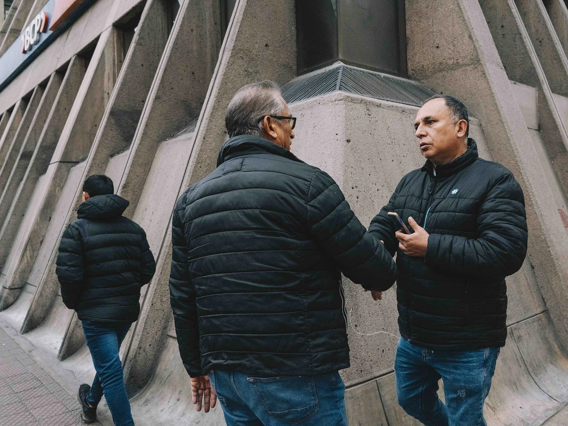 Two men stand facing each other and appear to be engaged in conversation, with one holding a phone, near a modern concrete building with angular architecture. In the background, another person walks away. All are dressed in dark jackets.