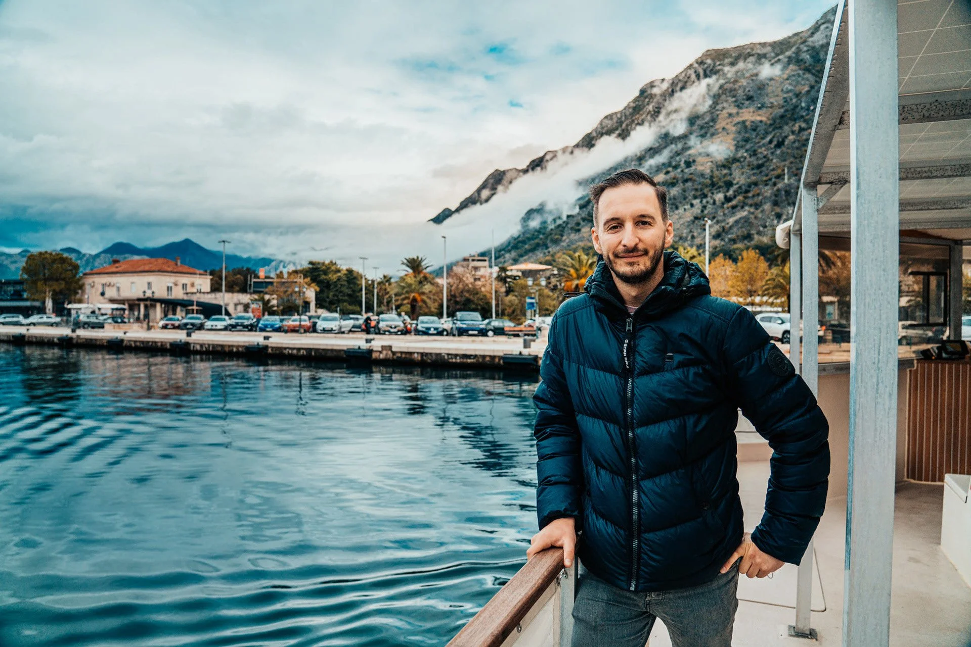 A man in a navy jacket standing on a boat dock, with a mountain and cloudy sky in the background.