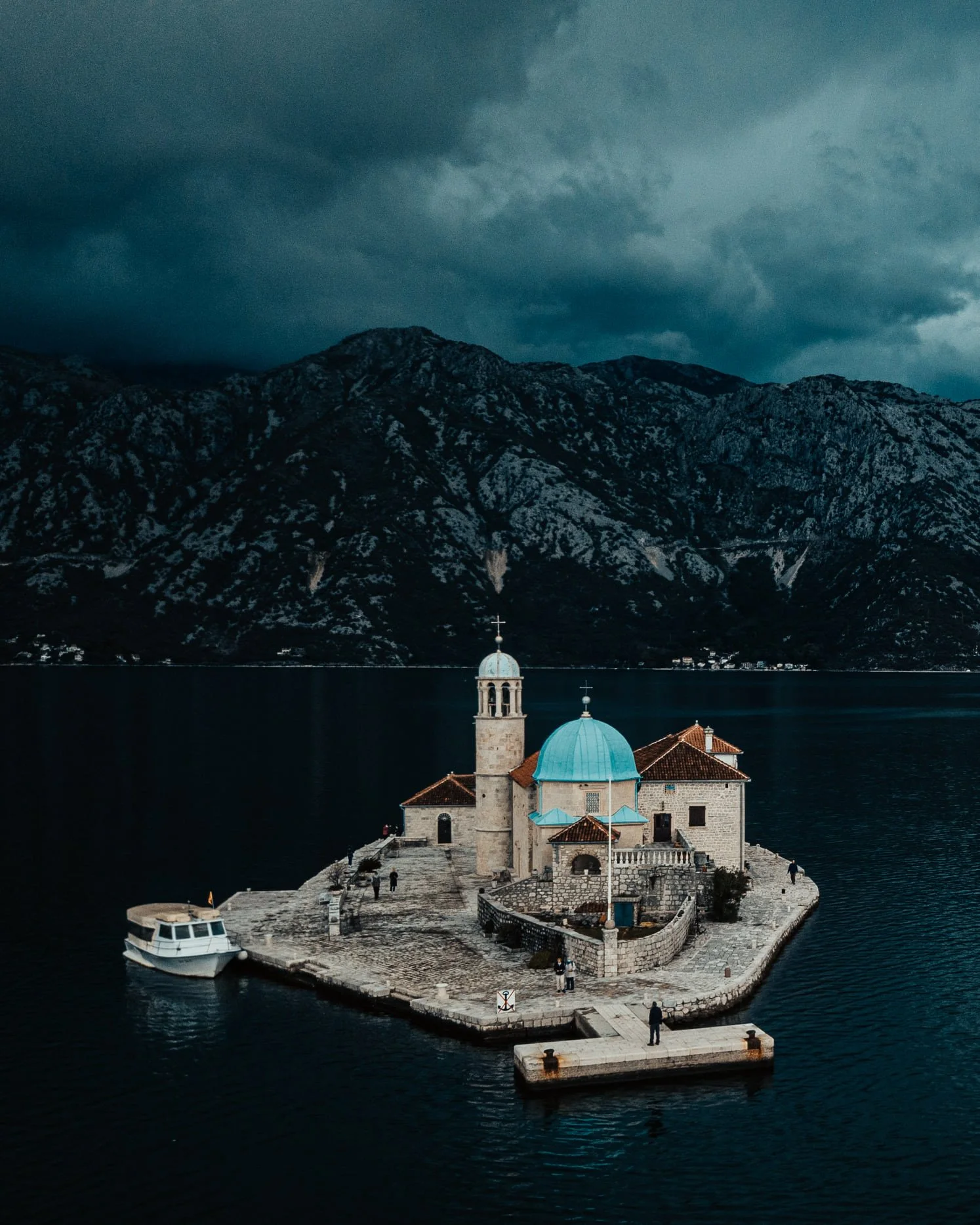A church on a small island with a blue dome, surrounded by water, with mountains and a dark, cloudy sky in the background.