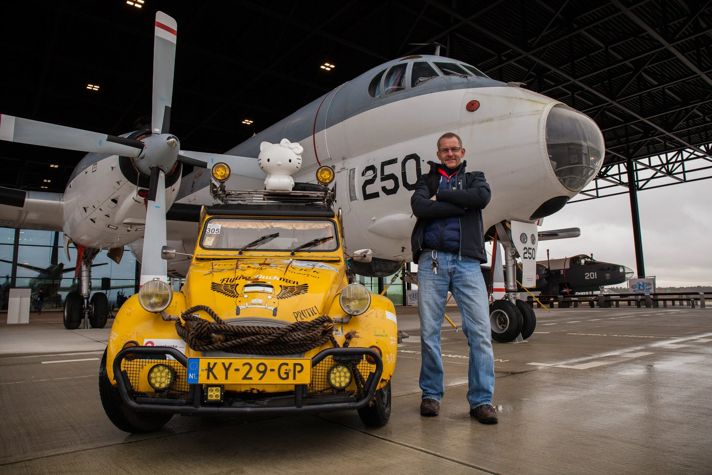 A man standing with arms crossed in front of a large military aircraft inside a hangar. In front of him is a yellow vehicle with a hello kitty figure on top. The aircraft and vehicle are on a tarmac.