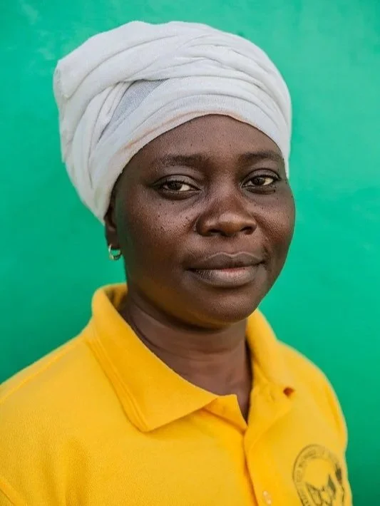 Woman wearing a white headwrap and yellow polo shirt standing against a green background.