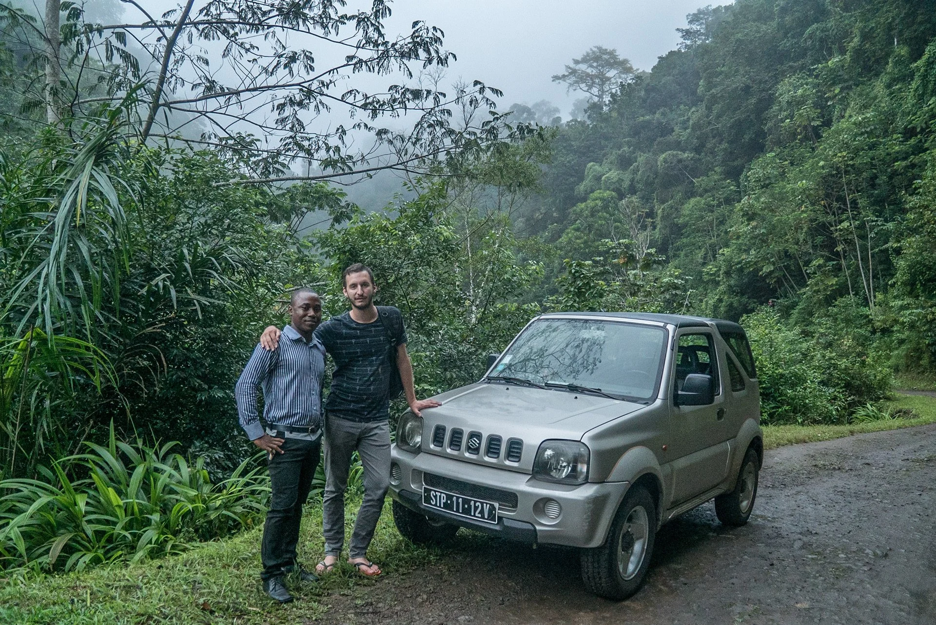 Two men standing next to a silver Suzuki vehicle on a dirt road surrounded by lush green jungle with mist and trees in the background.