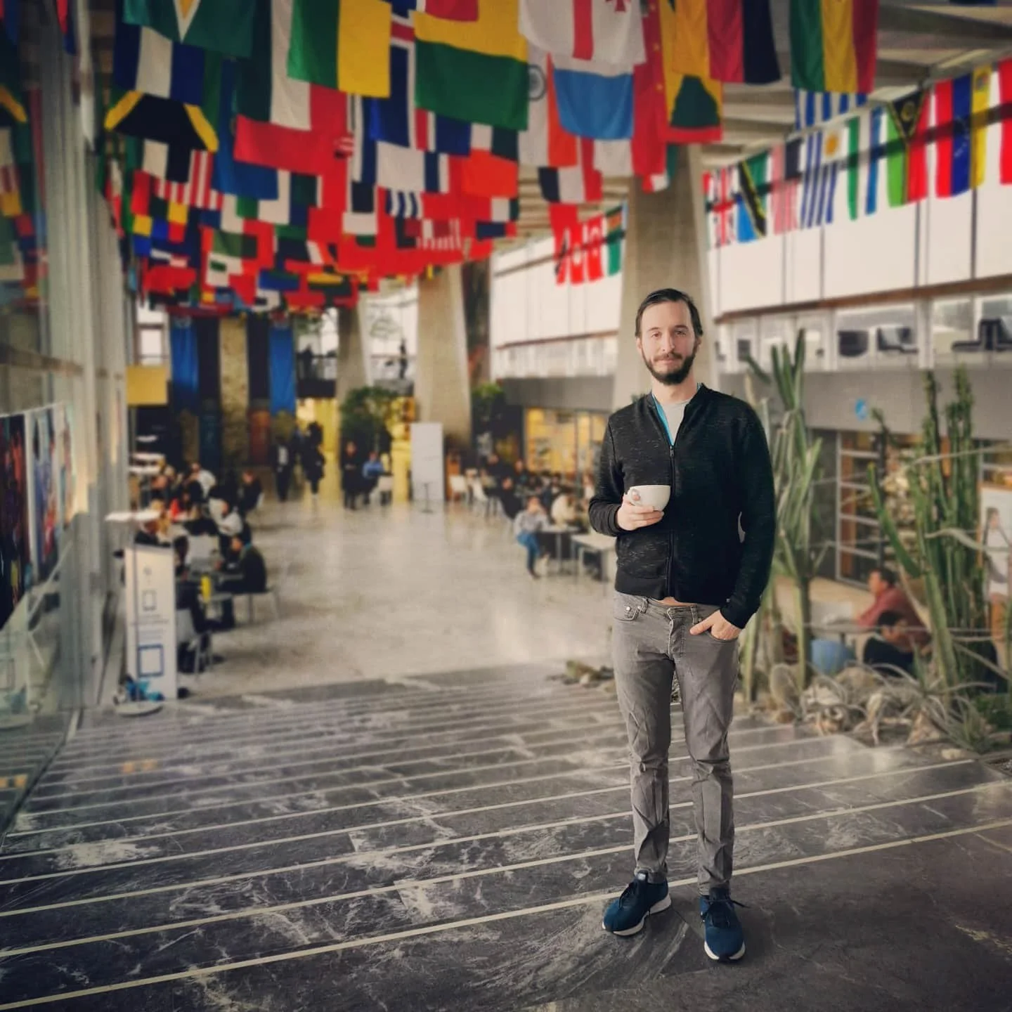 A man with a beard in a black jacket and gray pants holding a white mug, standing in an indoor public space decorated with colorful international flags hanging from the ceiling.