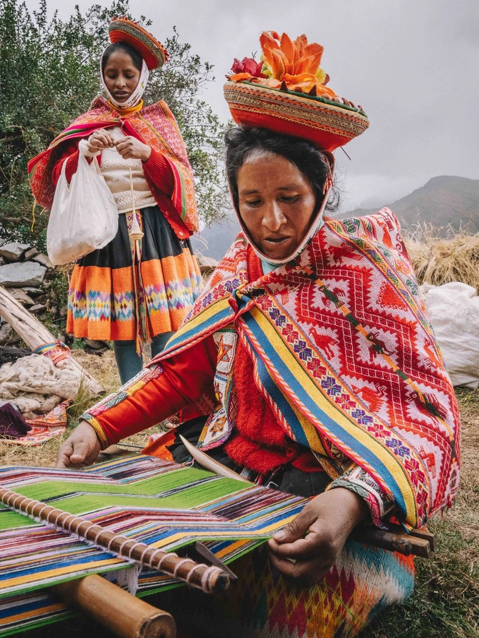 Two indigenous women in colorful traditional clothing, one sitting and working on a woven textile and the other standing, holding a bag, outdoors with mountains in the background.