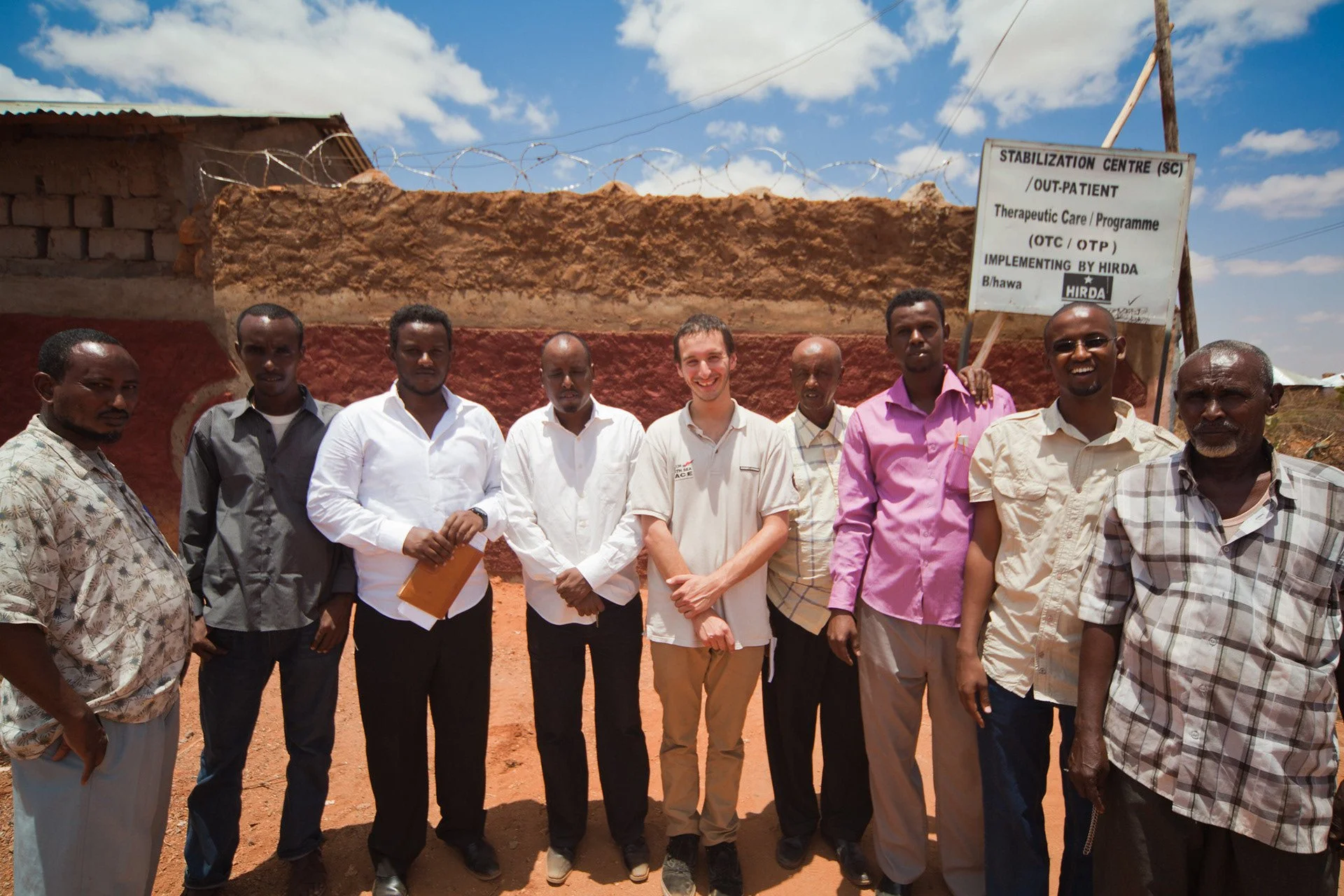 Group of people standing outdoors in front of a wall and barbed wire, smiling at the camera, with a sign indicating a stabilization center for therapeutic care in an African setting.