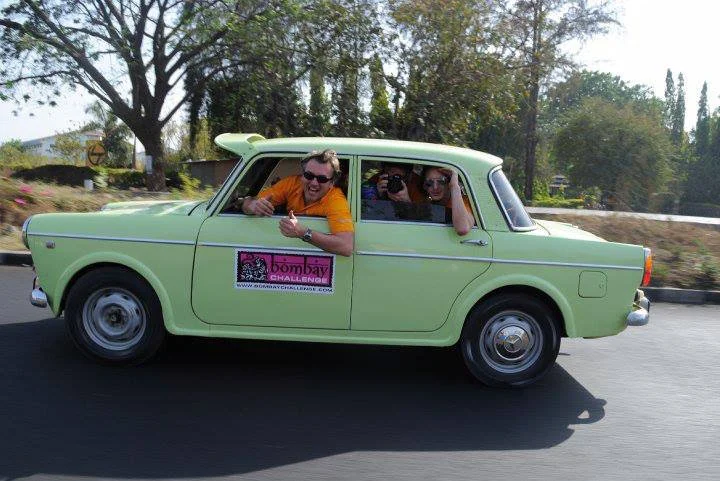 A vintage light green car with four people inside, two of whom are leaning out of the windows and smiling. The car has a pink sign on the side that says 'Bombay Challenge.' They are on a road with trees in the background on a sunny day.