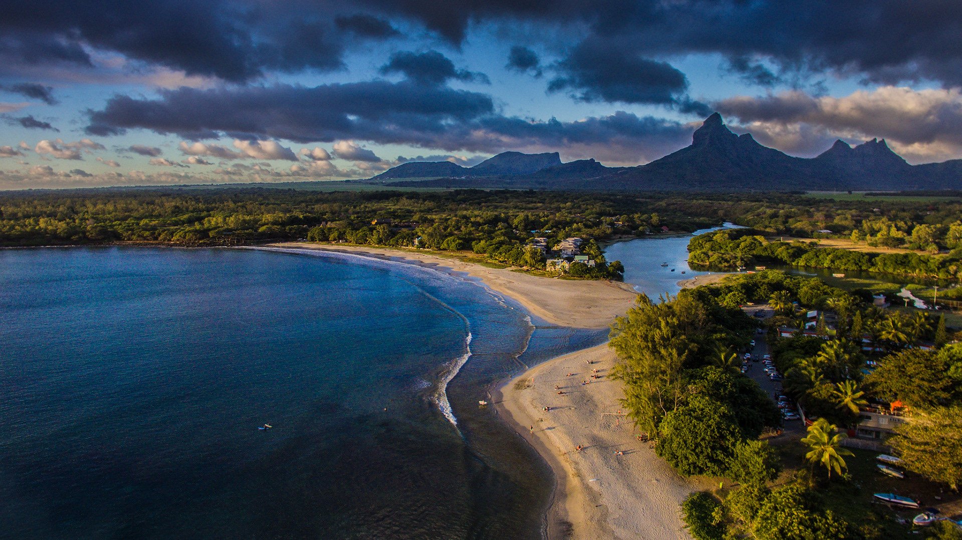 Aerial view of a tropical beach with a curved shoreline, calm blue ocean, lush greenery, and mountains in the distance under a partly cloudy sky.