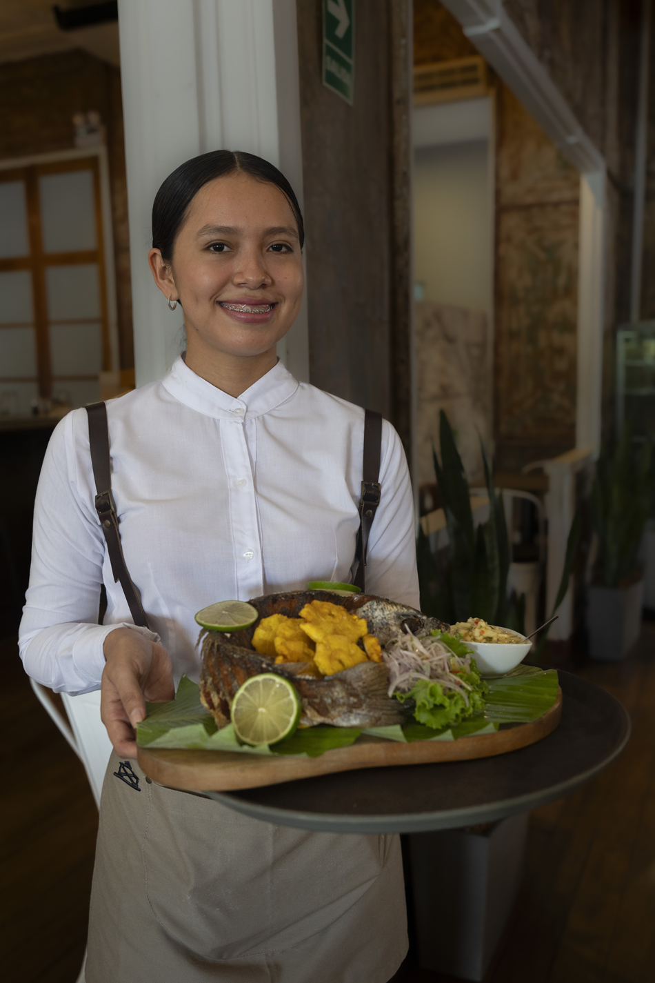 A smiling female server holding a wooden tray with a whole grilled fish topped with yellow sauce, lime wedges, a side of shredded vegetables with lettuce, and a small bowl of salad in a restaurant setting.