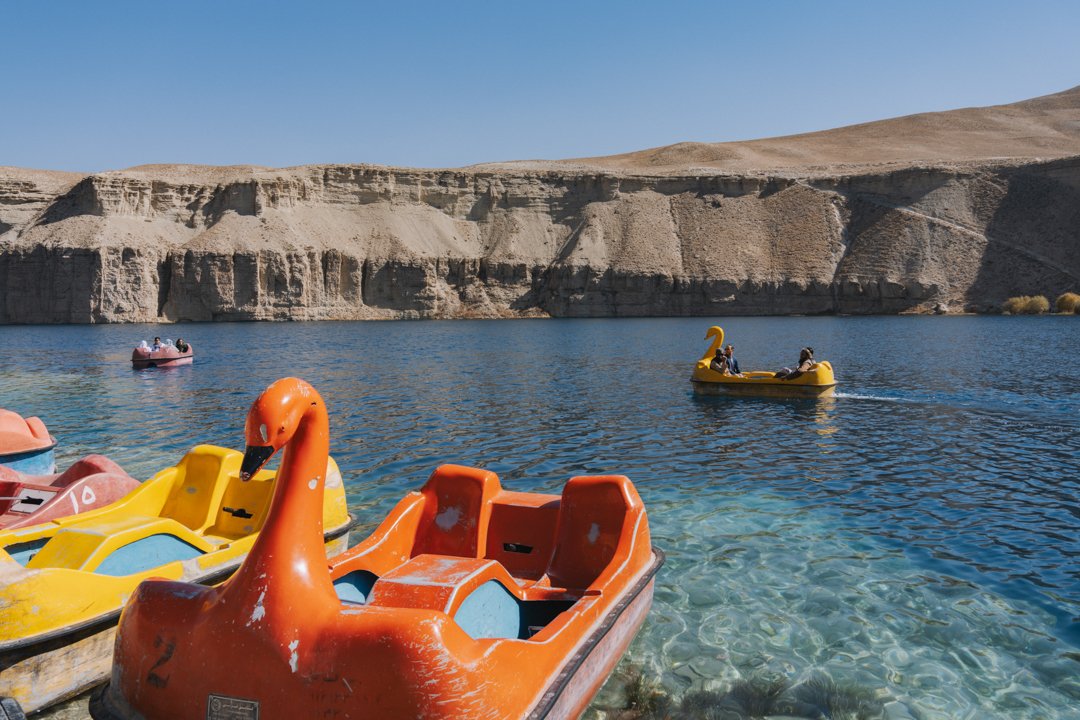 Colorful pedal boats shaped like swans and other animals floating on a clear lake with rocky cliffs in the background and a blue sky.