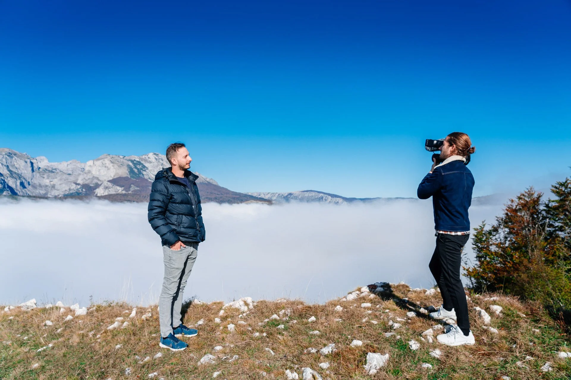 Two men stand on a grassy hilltop with rocky ground, the man on the right is taking a photo of the man on the left, both are wearing jackets, in the background are snow-capped mountains and a blue sky, with a layer of fog in the valley below.