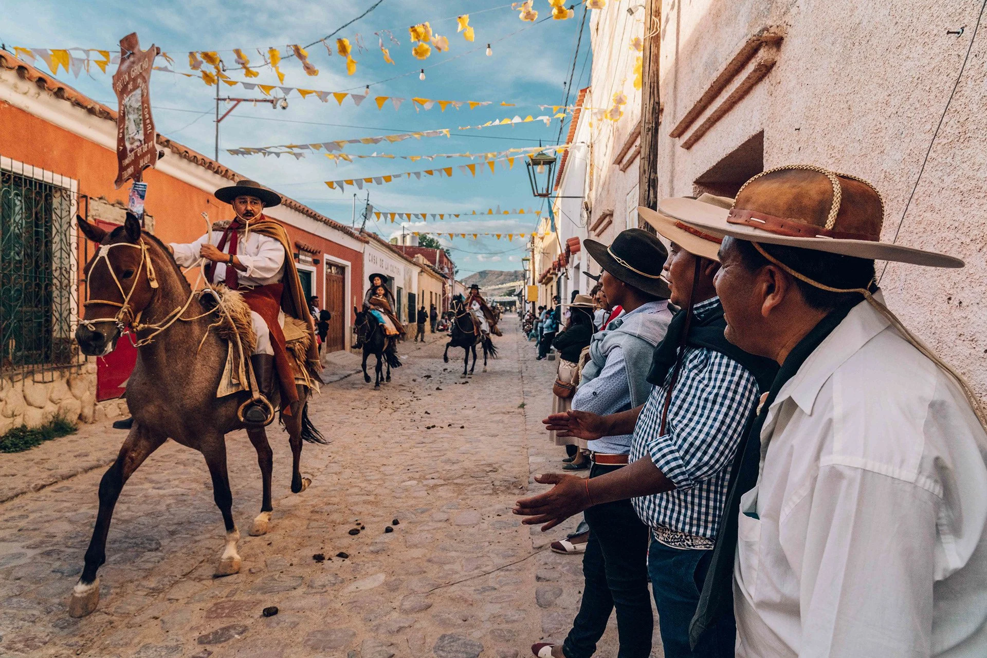 Street scene with people watching a traditional horse parade, colorful banners and buildings with vibrant colors, clear sky.