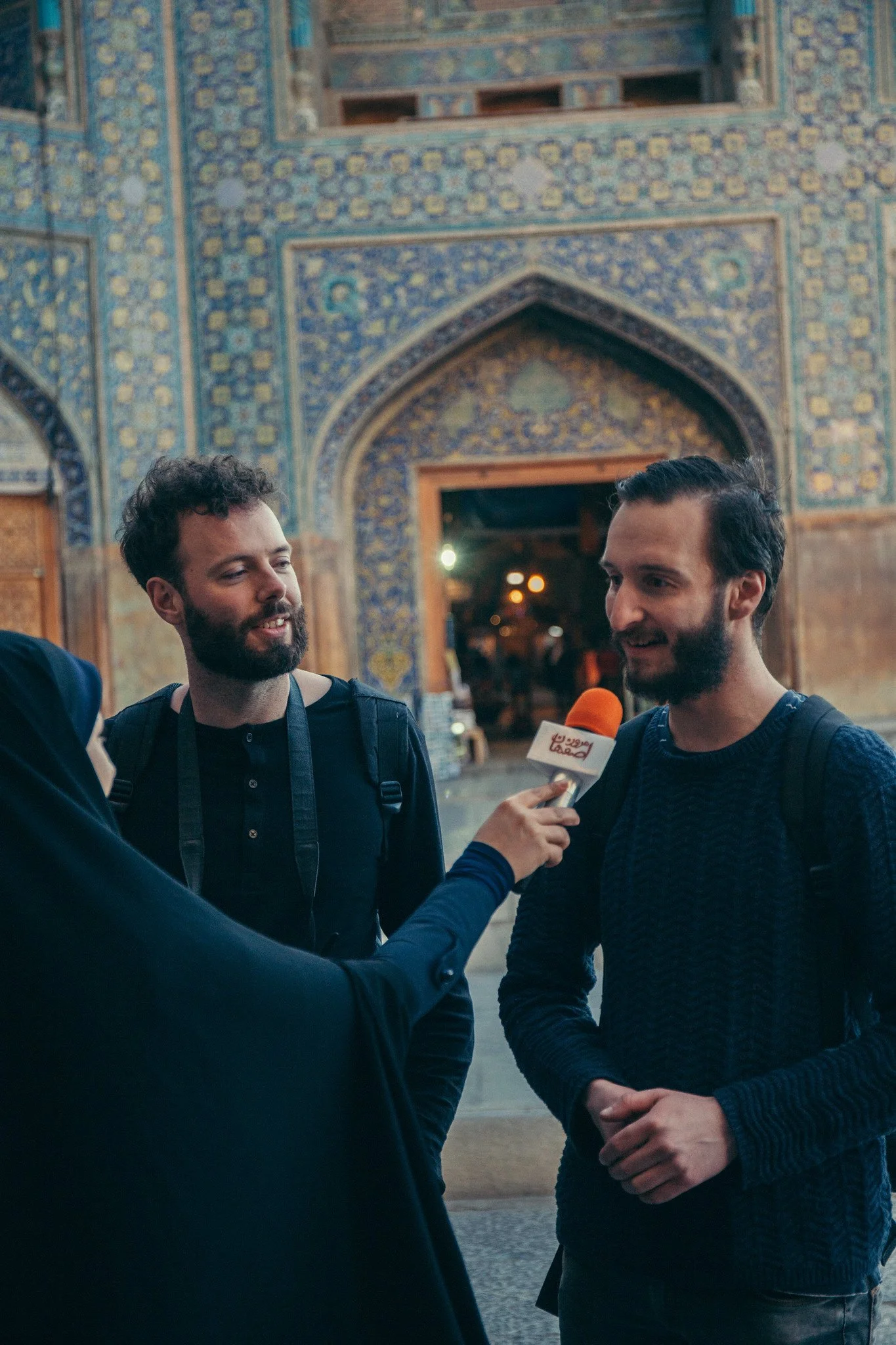 Two men are being interviewed in front of a historic building with intricate blue and yellow tilework and arched doorways. The interviewer is holding a microphone with a logo and is dressed in a dark blazer, while the interviewees are casually dressed, smiling, and appear engaged in conversation.