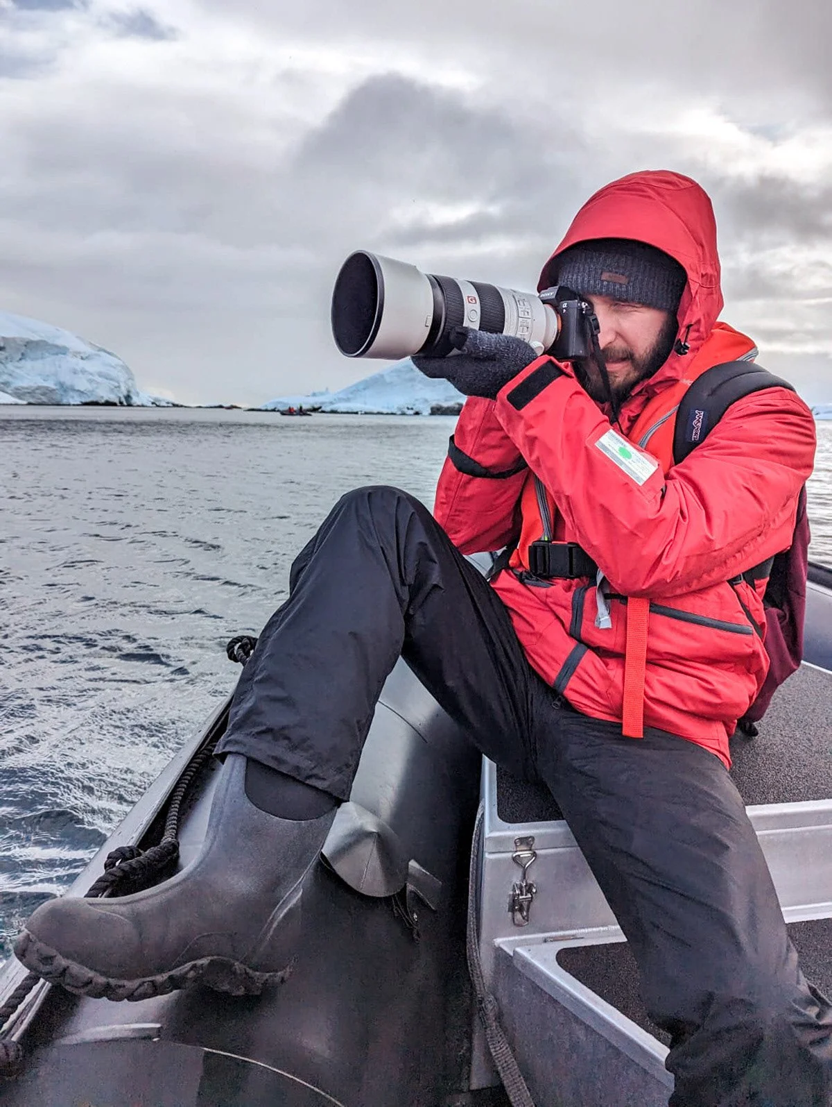 A man dressed in red winter gear and black pants, sitting on a boat, taking photos with a large telephoto lens camera. He is wearing gloves, a hat, and rubber boots, with icy landscape and icebergs in the background.