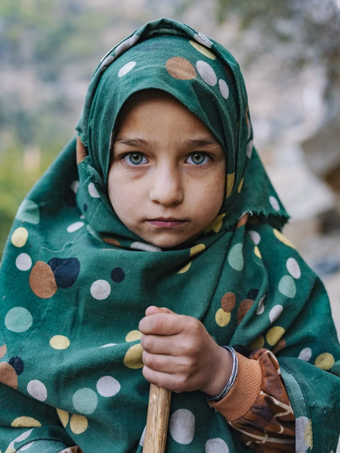A young child with blue eyes wearing a polka-dot headscarf, holding a stick, outdoors with a blurred natural background.