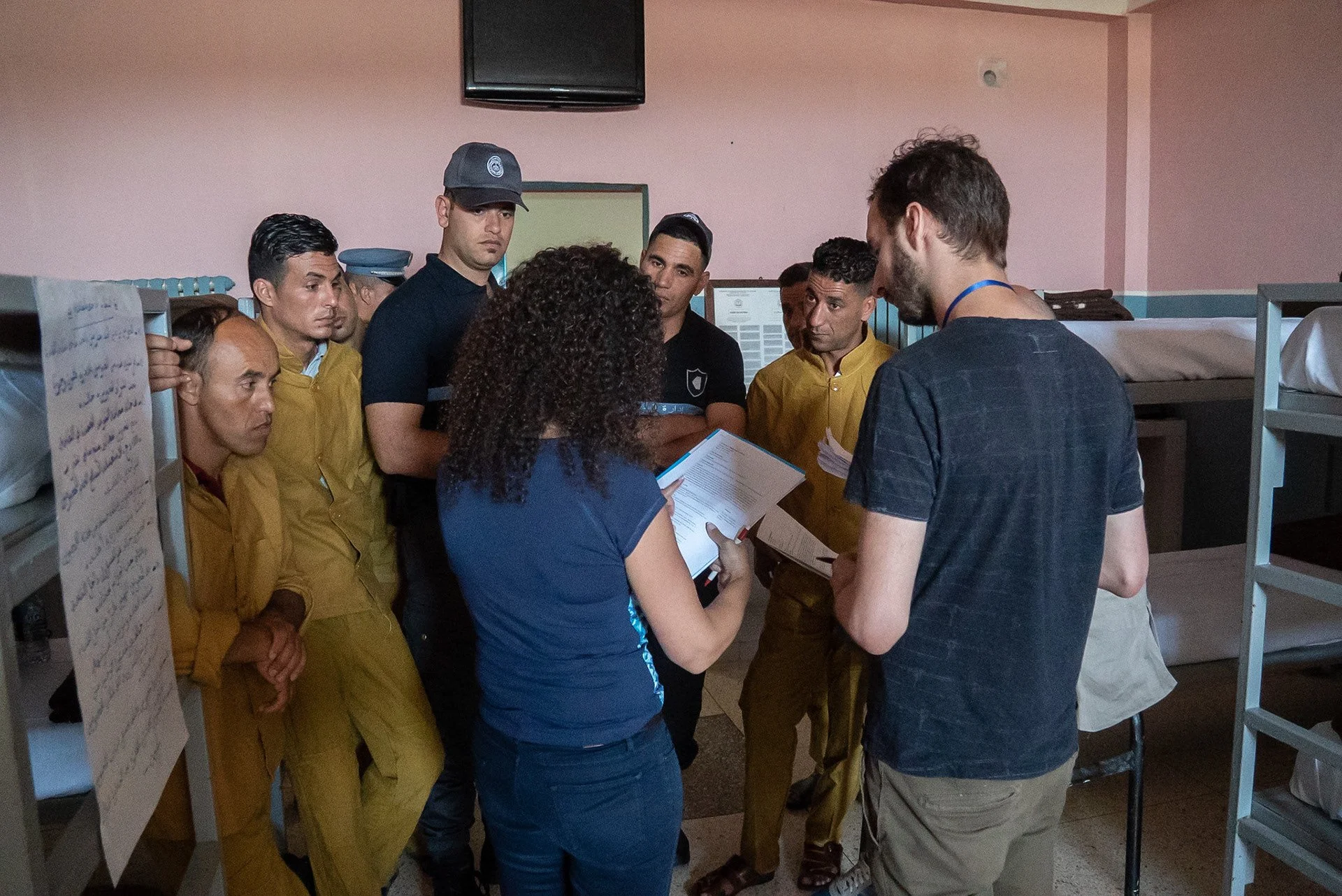 Group of people, some in yellow uniforms, gathered around a woman holding a clipboard, in what appears to be an indoor setting with bunk beds and a pink wall.